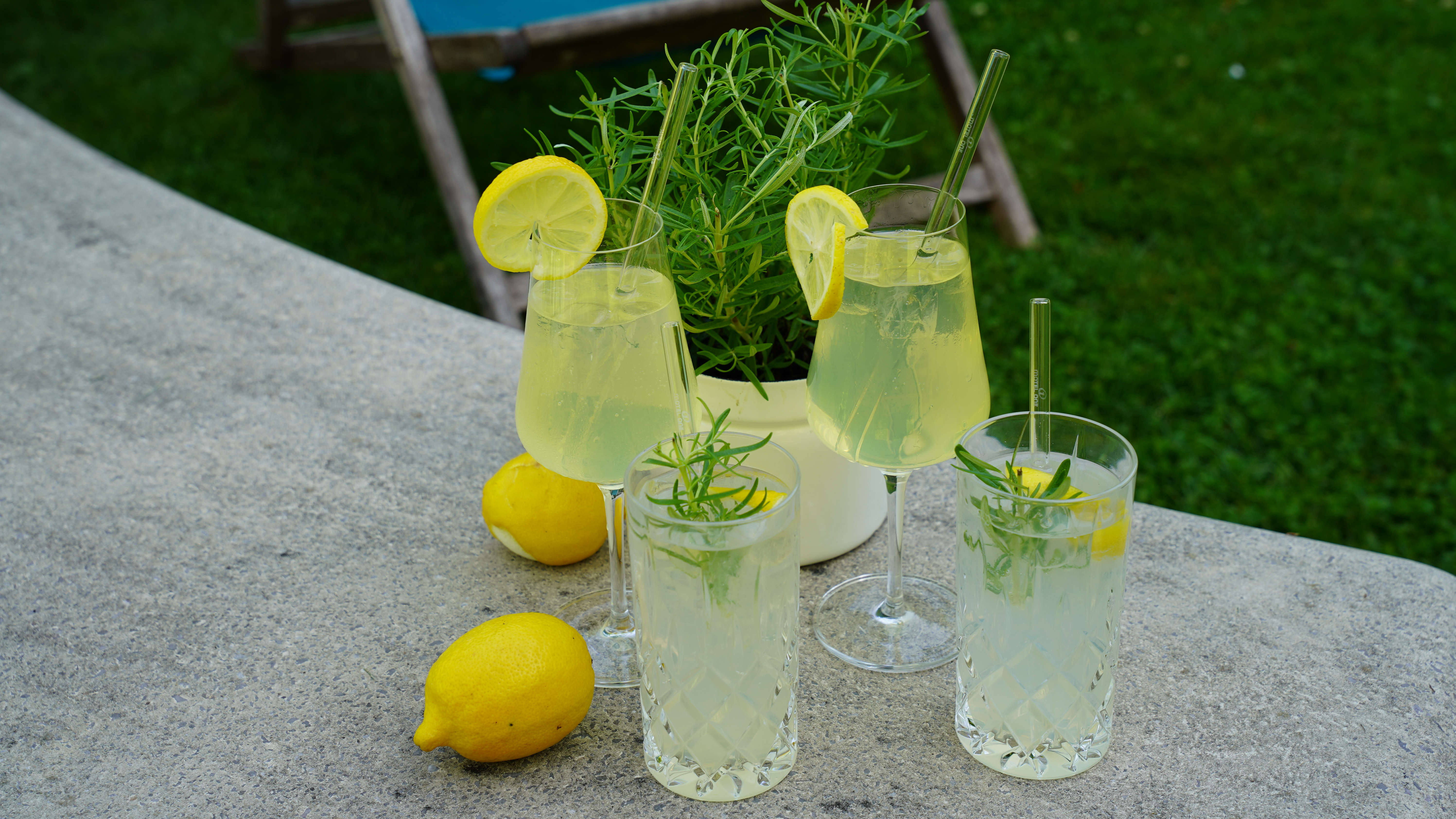 Frische Zitronenlimonade mit Kräutern in Gläsern auf Tisch, umgeben von Zitronen und einem Kräutertopf, entspannte Gartenszenerie mit Liegestuhl im Hintergrund.