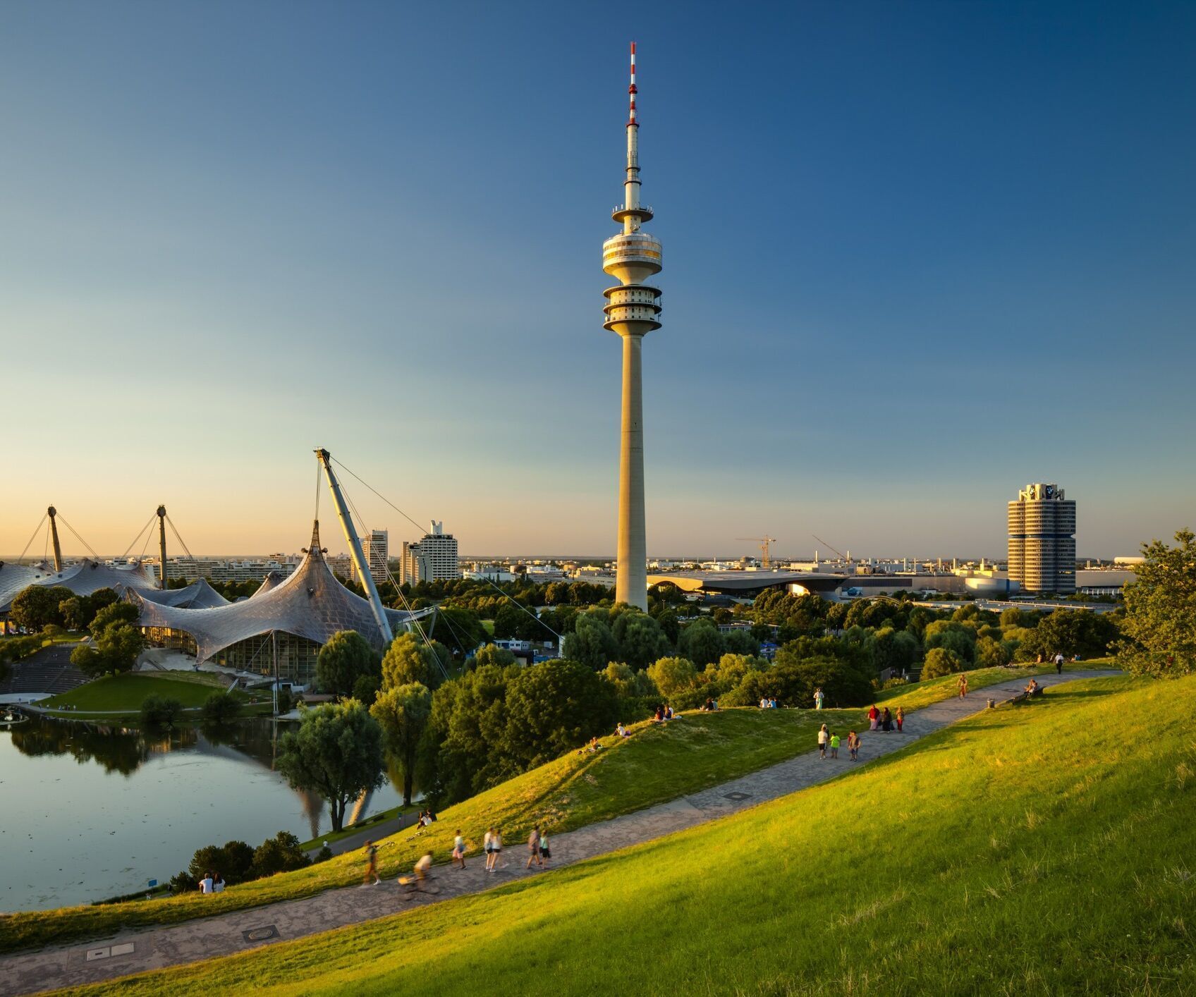 Blick auf den Olympiapark in München bei Sonnenuntergang, mit dem Olympiaturm, einem See, grünen Hügeln und Menschen, die den Weg entlang spazieren.