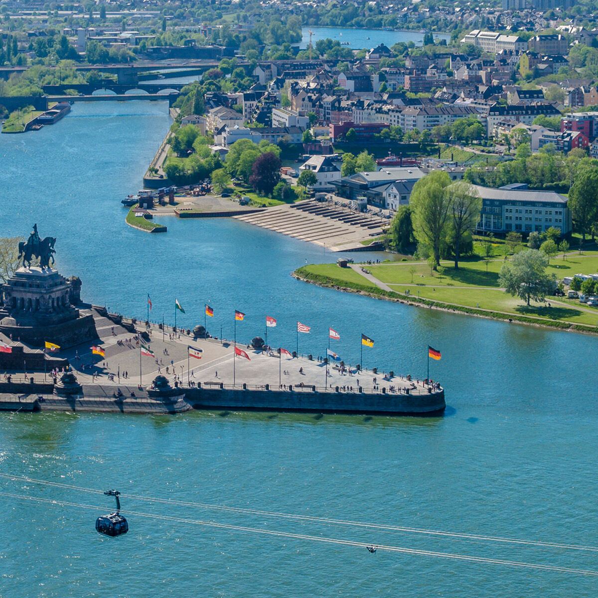 Luftaufnahme eines Flussufers mit einer markanten Statue auf einer Halbinsel, mehreren Brücken im Hintergrund und einem Schiff auf dem Wasser, umgeben von einer grünen, städtischen Landschaft unter klarem Himmel.
