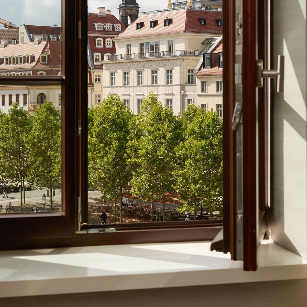 Blick aus einem geöffneten Fenster auf einen belebten Stadtplatz mit grünen Bäumen und historischen Gebäuden im Hintergrund unter bewölktem Himmel.