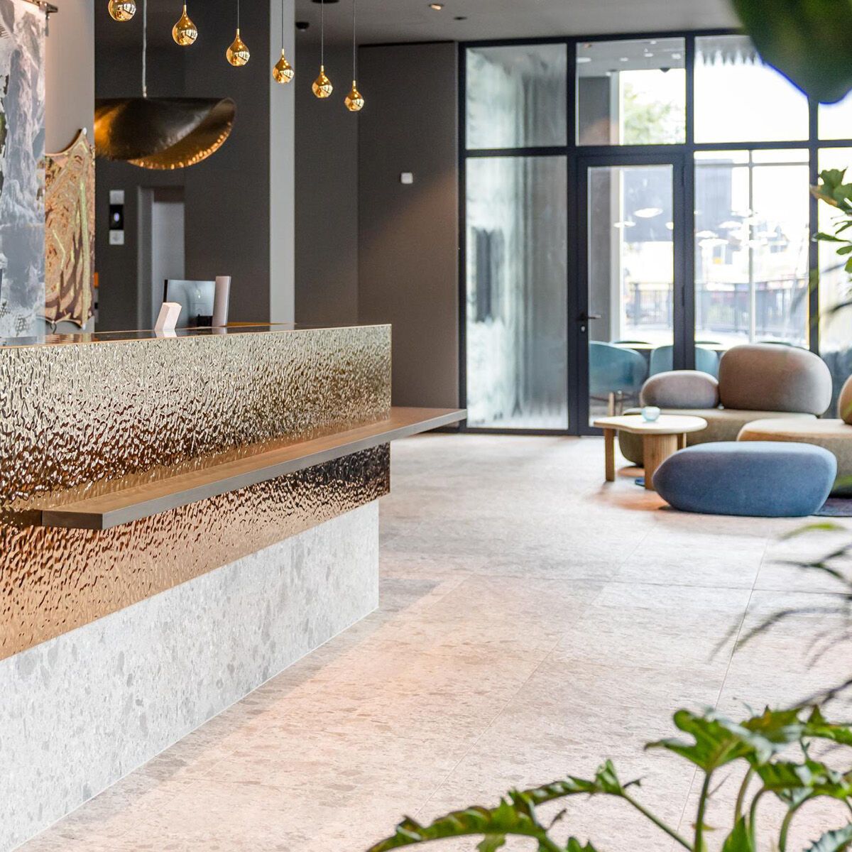 Moderner Hotellobby-Empfang mit elegantem, goldfarbenem Tresen, stilvollen Pendelleuchten und gemütlichen Sitzbereichen mit großen Fenstern für viel Tageslicht. Einladende Atmosphäre für Geschäfts- und Urlaubsreisende.