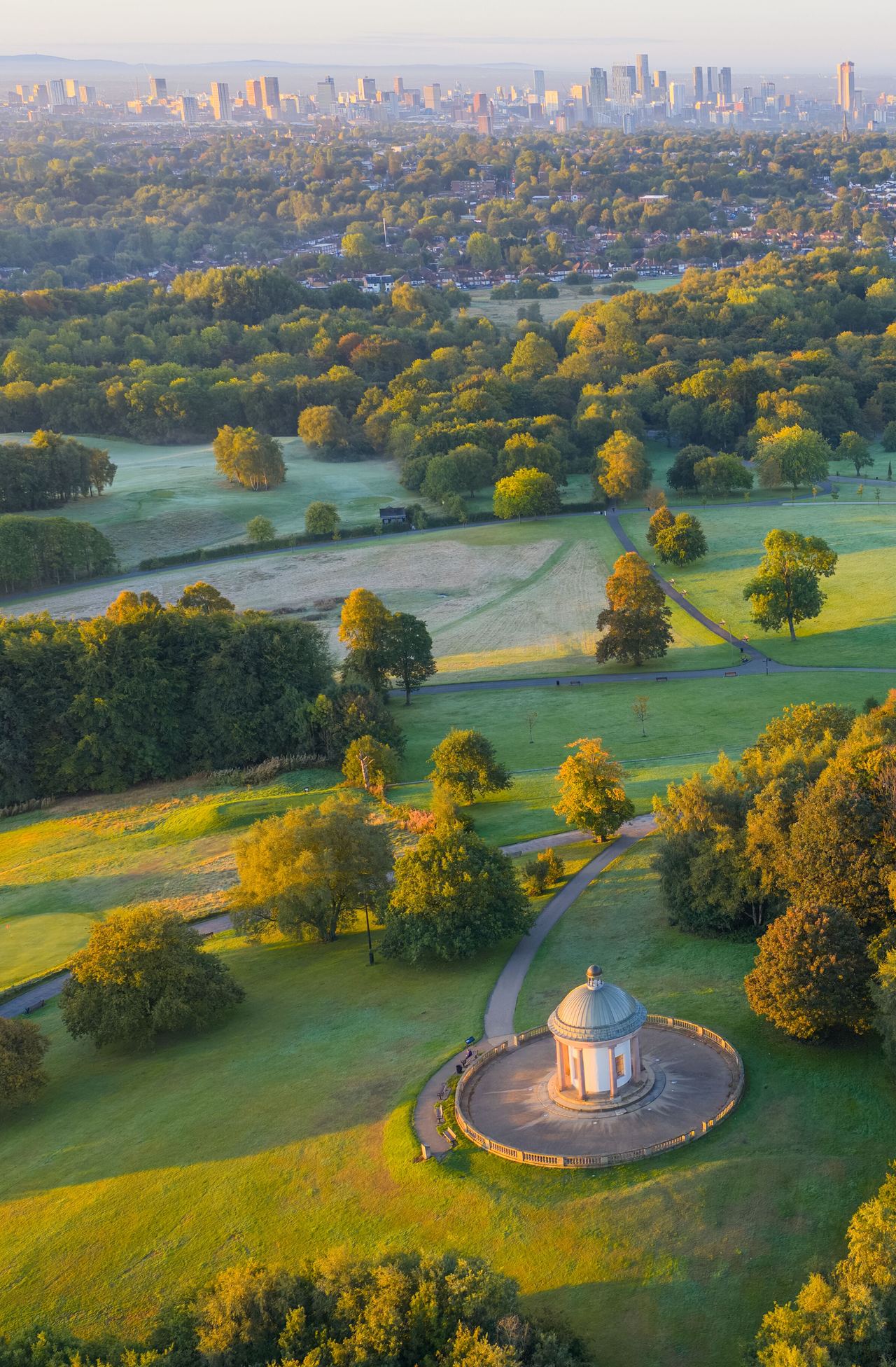 Weitblick auf einen weitläufigen Park mit grünen Wiesen und herbstlich gefärbten Bäumen, einem historischen Pavillon und einem großen Herrenhaus, im Hintergrund die Skyline einer Stadt unter blauem Himmel.