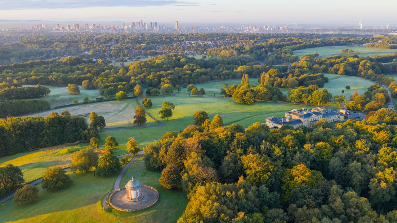 Weitblick auf einen weitläufigen Park mit grünen Wiesen und herbstlich gefärbten Bäumen, einem historischen Pavillon und einem großen Herrenhaus, im Hintergrund die Skyline einer Stadt unter blauem Himmel. Weitblick auf einen weitläufigen Park mit grünen Wiesen und herbstlich gefärbten Bäumen, einem historischen Pavillon und einem großen Herrenhaus, im Hintergrund die Skyline einer Stadt unter blauem Himmel.