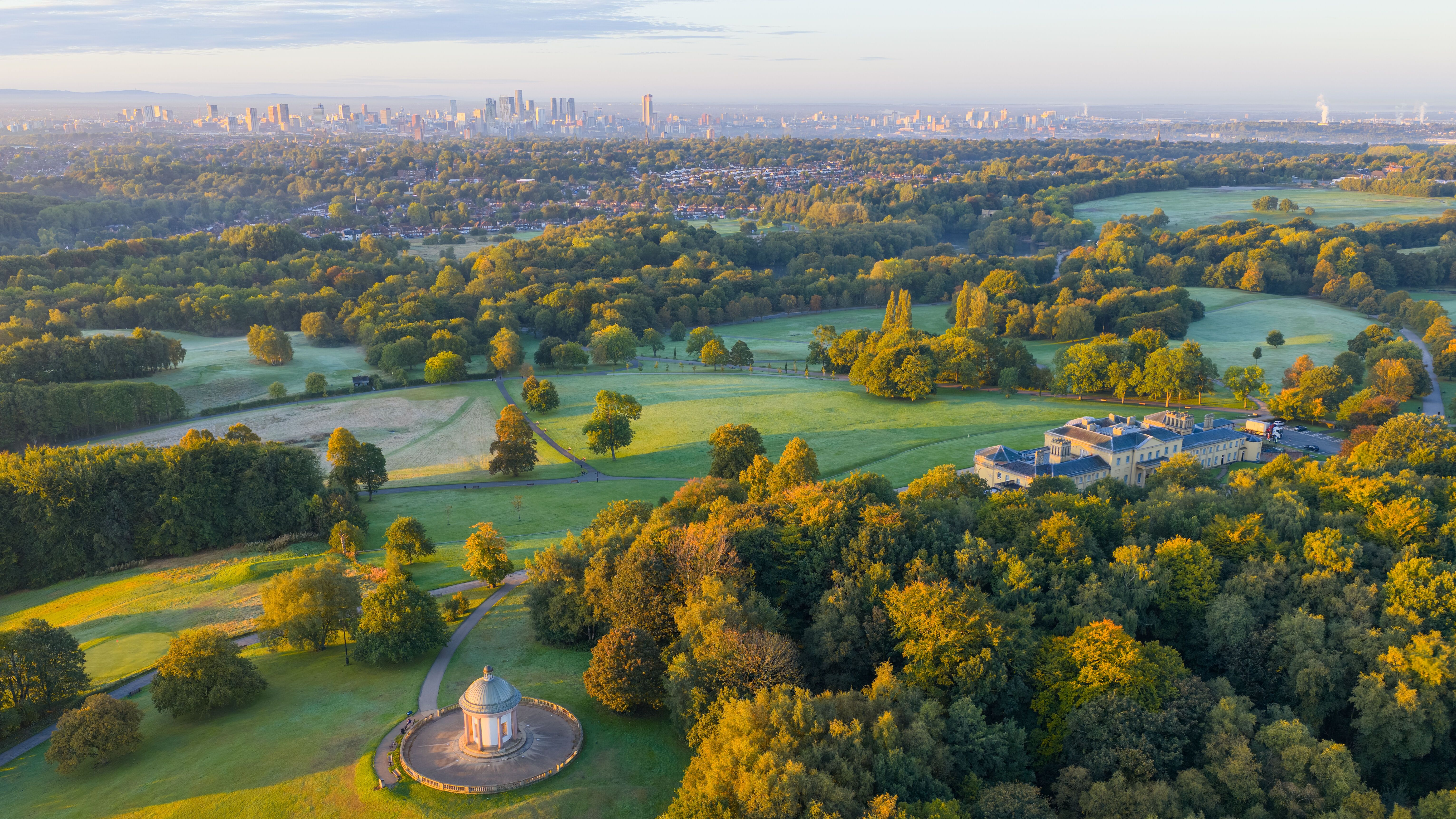 Weitblick auf einen weitläufigen Park mit grünen Wiesen und herbstlich gefärbten Bäumen, einem historischen Pavillon und einem großen Herrenhaus, im Hintergrund die Skyline einer Stadt unter blauem Himmel.