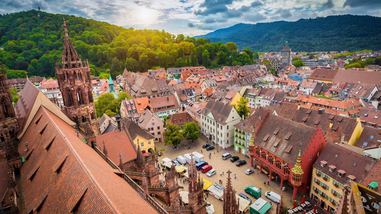 Blick über die historische Altstadt Freiburg mit Fachwerkhäusern, gepflastertem Marktplatz und einer markanten gotischen Kirche, umgeben von bewaldeten Hügeln und Bergen im Sonnenlicht.