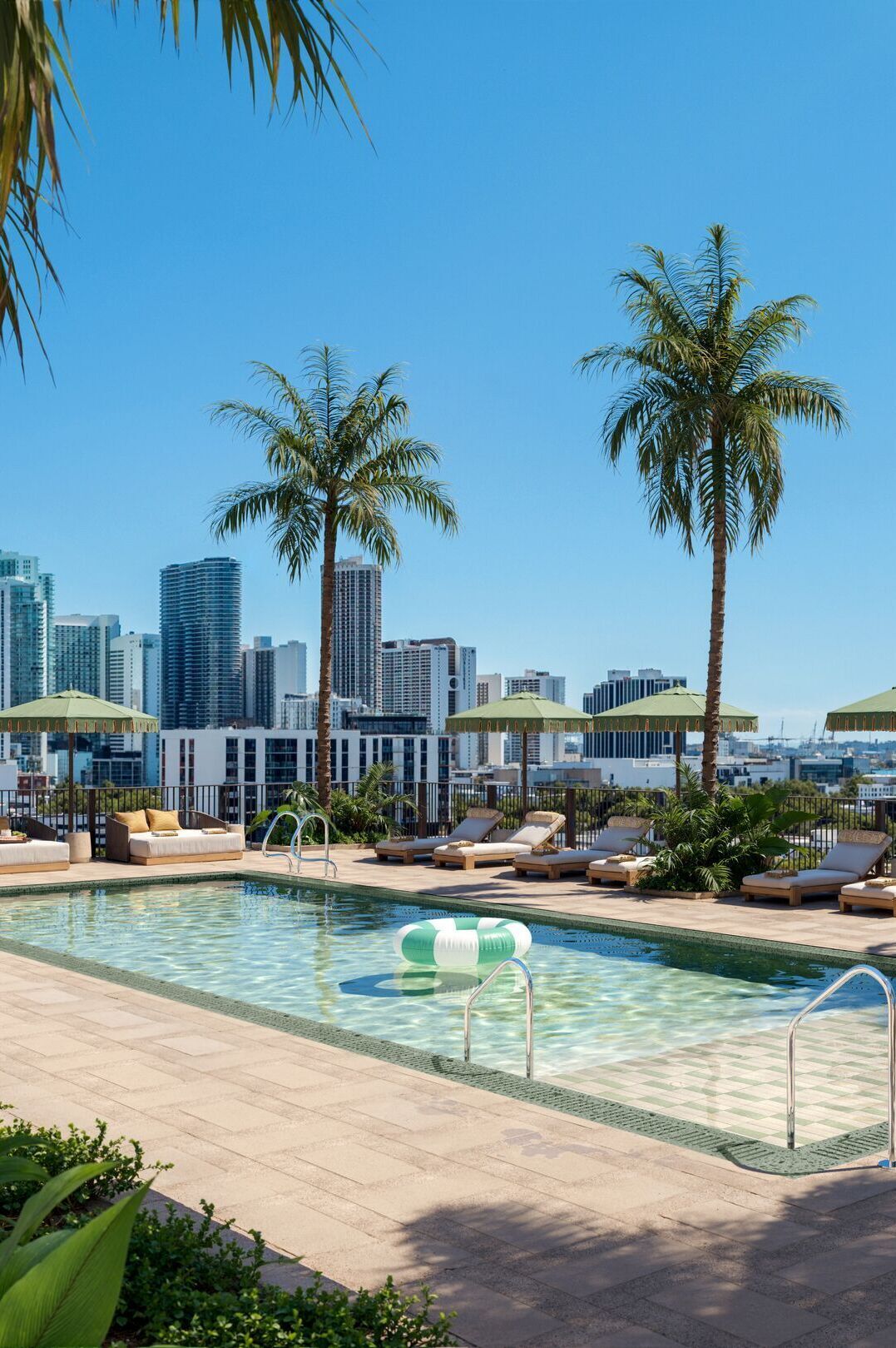 Dachterrassen-Pool mit Liegestühlen und Sonnenschirmen, umgeben von Palmen, mit Blick auf moderne Hochhäuser und blauen Himmel – ideal für Entspannung und urbanen Freizeitgenuss.