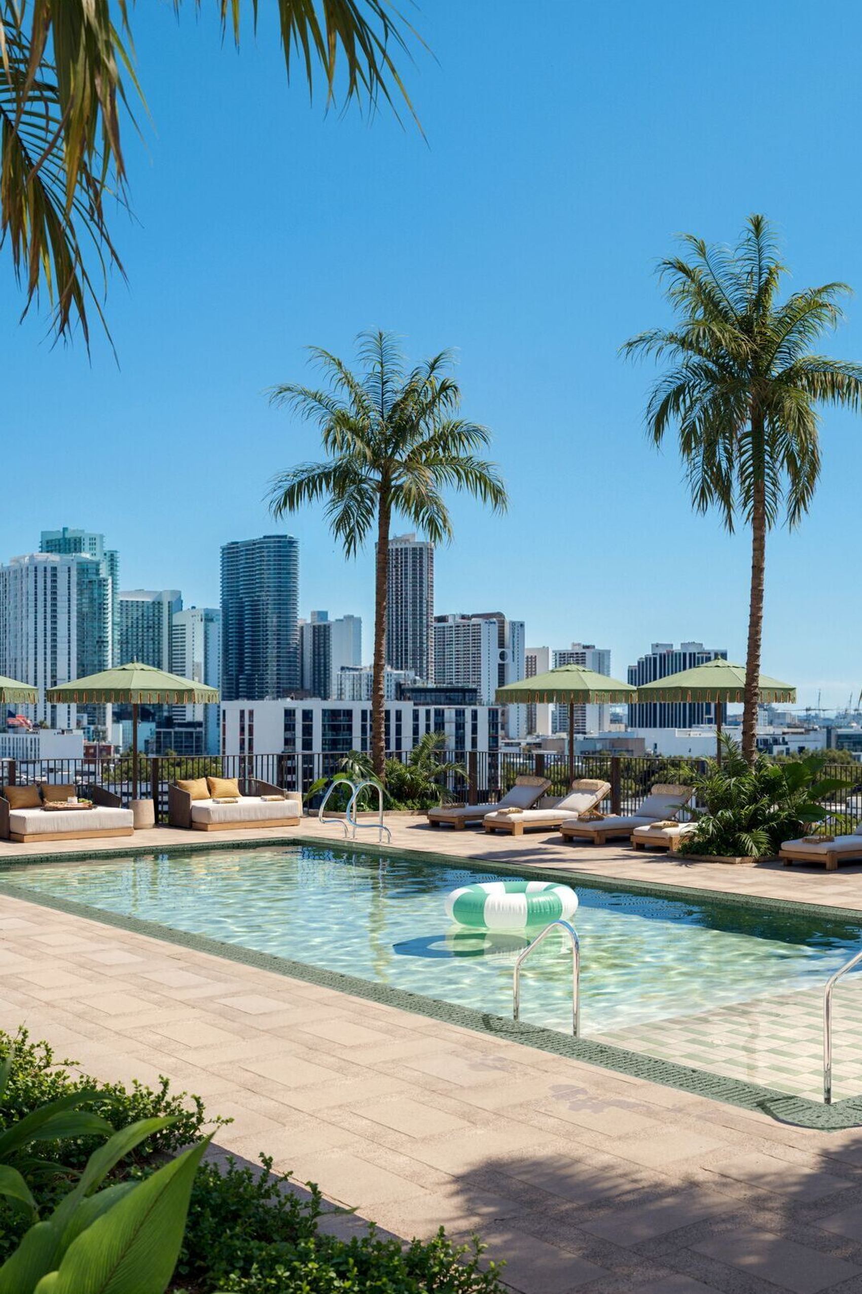 Dachterrassen-Pool mit Liegestühlen und Sonnenschirmen, umgeben von Palmen, mit Blick auf moderne Hochhäuser und blauen Himmel – ideal für Entspannung und urbanen Freizeitgenuss.