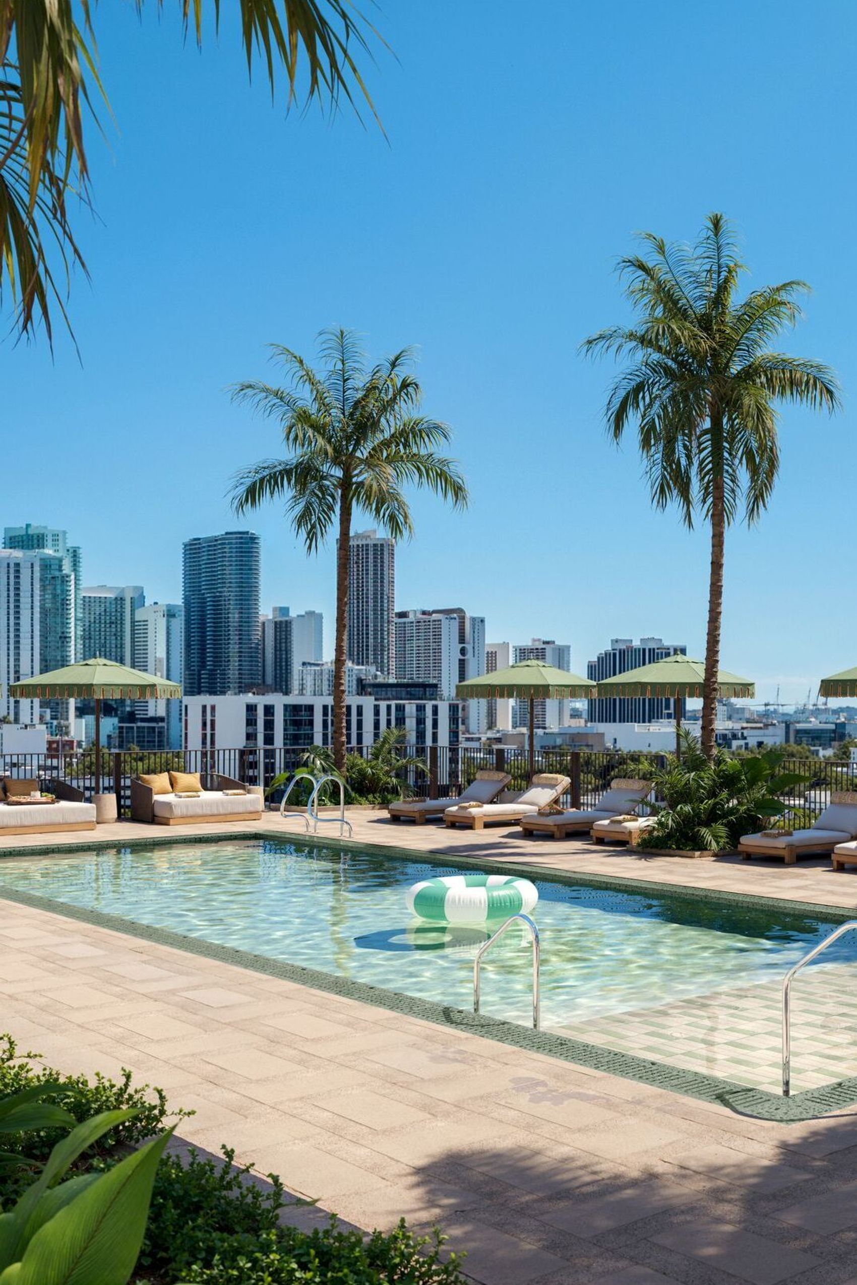 Dachterrassen-Pool mit Liegestühlen und Sonnenschirmen, umgeben von Palmen, mit Blick auf moderne Hochhäuser und blauen Himmel – ideal für Entspannung und urbanen Freizeitgenuss.