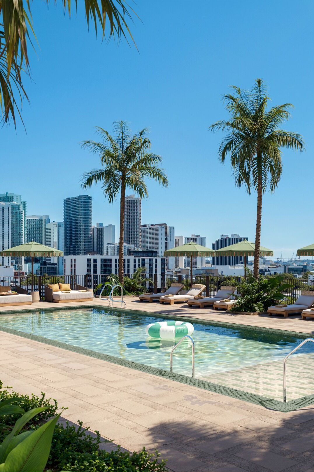 Dachterrassen-Pool mit Liegestühlen und Sonnenschirmen, umgeben von Palmen, mit Blick auf moderne Hochhäuser und blauen Himmel – ideal für Entspannung und urbanen Freizeitgenuss.