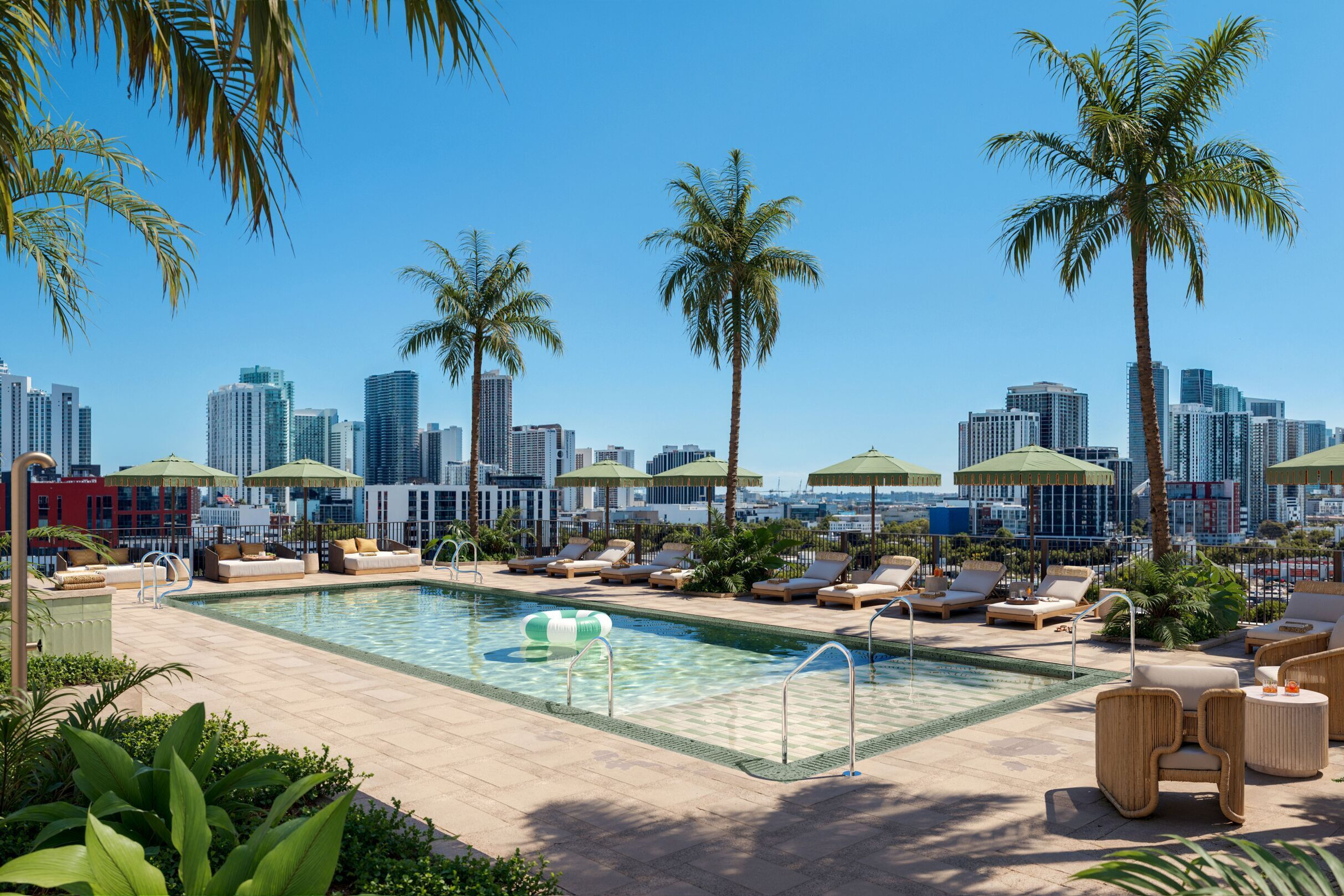Dachterrassen-Pool mit Liegestühlen und Sonnenschirmen, umgeben von Palmen, mit Blick auf moderne Hochhäuser und blauen Himmel – ideal für Entspannung und urbanen Freizeitgenuss.