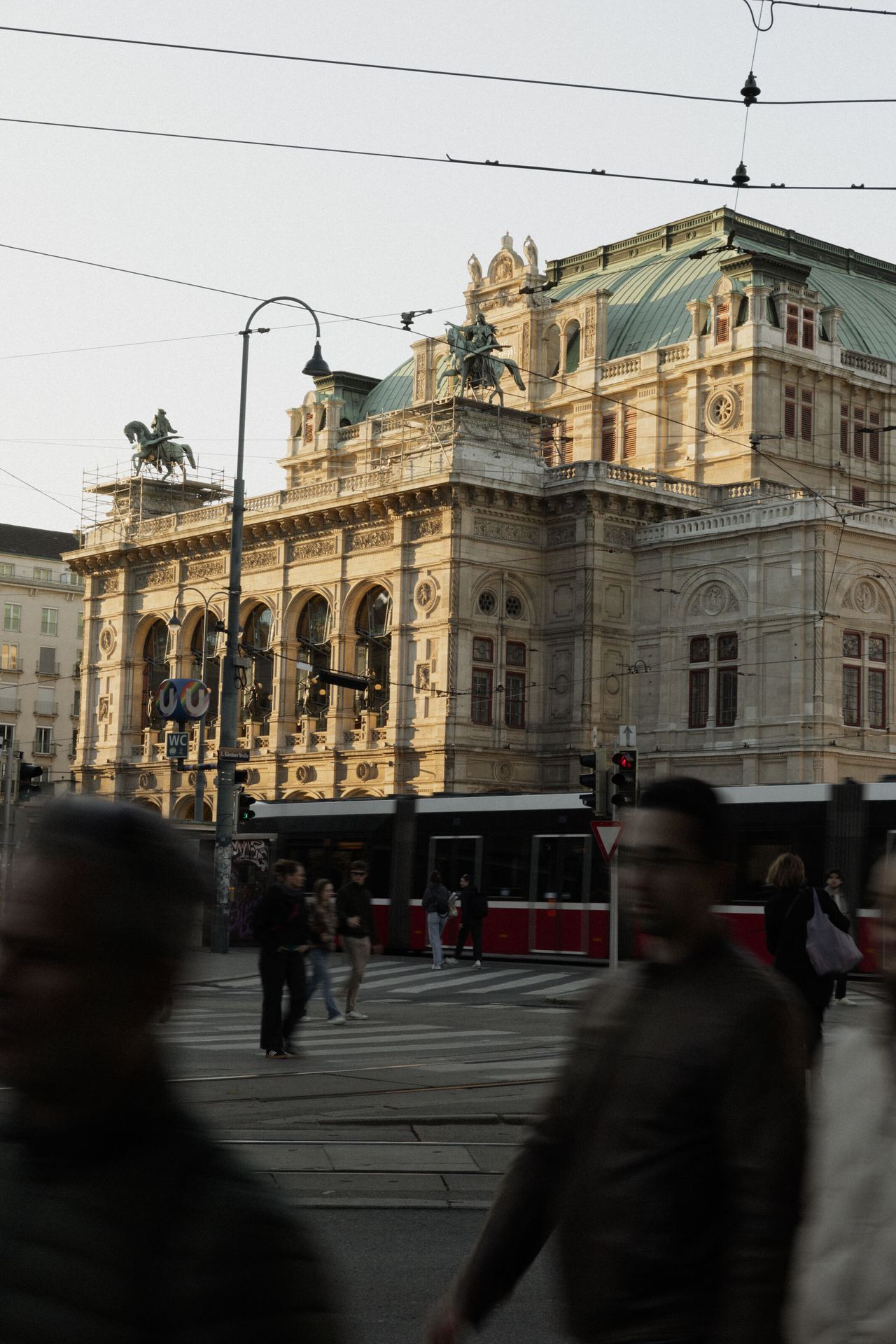 Historisches prunkvolles Theatergebäude mit aufwendiger Fassade und Statuen auf dem Dach, beleuchtet vom warmen Nachmittagslicht, im Vordergrund Straßenbahnen und Fußgänger an einer belebten Kreuzung.