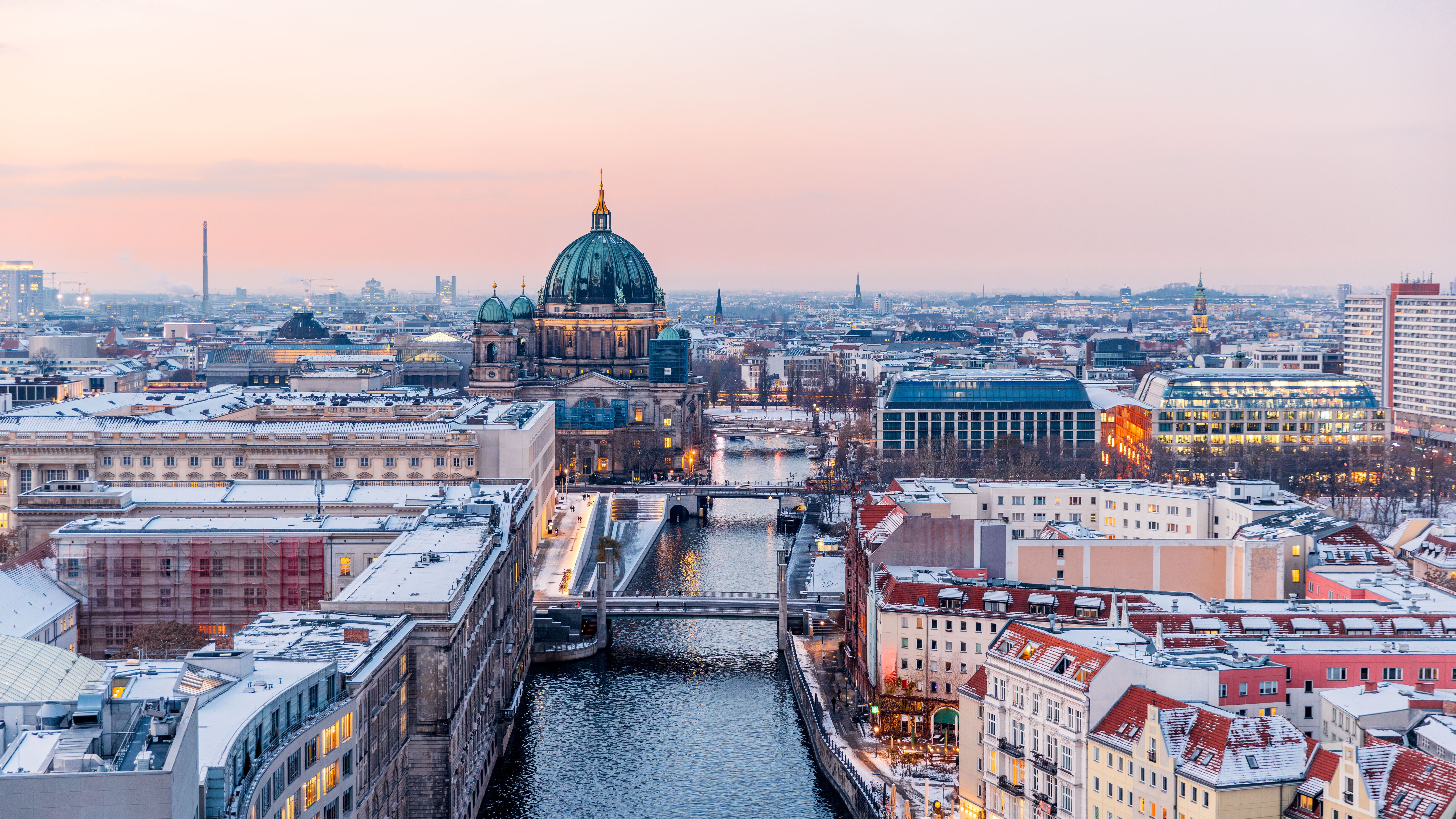 Blick auf die Berliner Stadtkulisse bei Dämmerung mit dem beleuchteten Berliner Dom und der Spree, die sich durch die Stadt schlängelt. Historische und moderne Gebäude prägen das Bild, das eine einladende Atmosphäre für Städtereisende und Kulturinteressierte vermittelt.