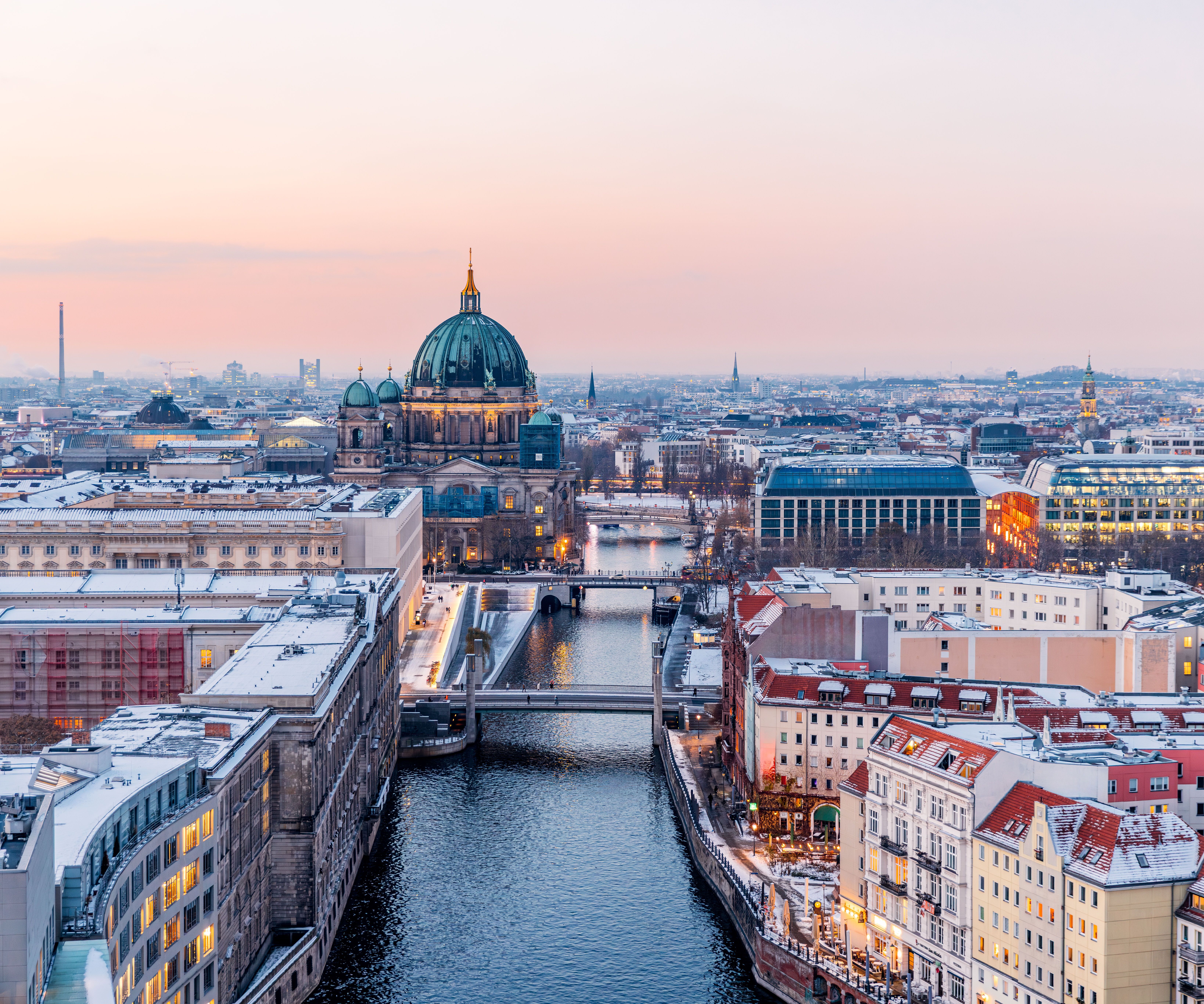 Blick auf die Berliner Stadtkulisse bei Dämmerung mit dem beleuchteten Berliner Dom und der Spree, die sich durch die Stadt schlängelt. Historische und moderne Gebäude prägen das Bild, das eine einladende Atmosphäre für Städtereisende und Kulturinteressierte vermittelt.