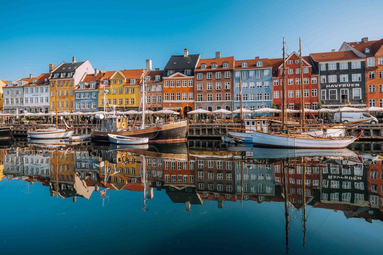 Bunte, historische Häuser säumen einen ruhigen Hafen mit festgemachten Segelbooten, die sich im klaren Wasser spiegeln, unter strahlend blauem Himmel.
