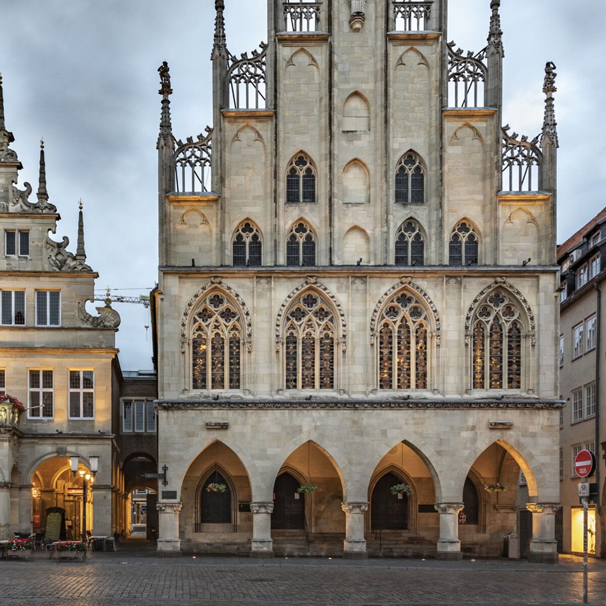 Historische gotische und renaissanceartige Gebäude mit detailreichen Fassaden und spitzen Türmen in einem belebten Stadtplatz unter wolkigem Himmel. Beleuchtete Arkaden laden zum Verweilen ein.