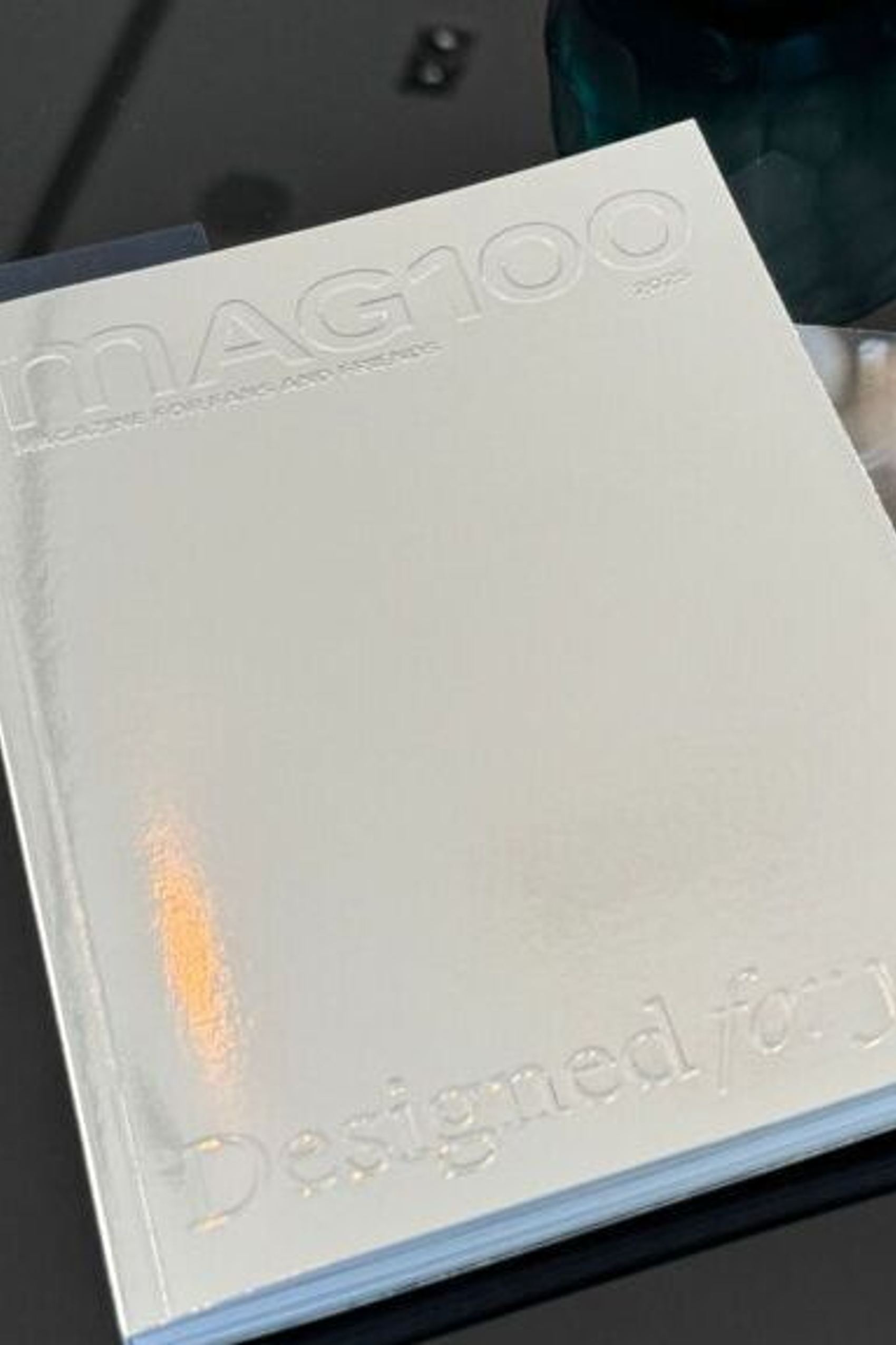 Weiße Broschüre mit dem Titel „MAG100“ und einem Werbeflyer mit lächelnder Frau, der auf Ersparnisse bei Hotelbuchungen hinweist, auf einem schwarzen runden Tisch liegend.