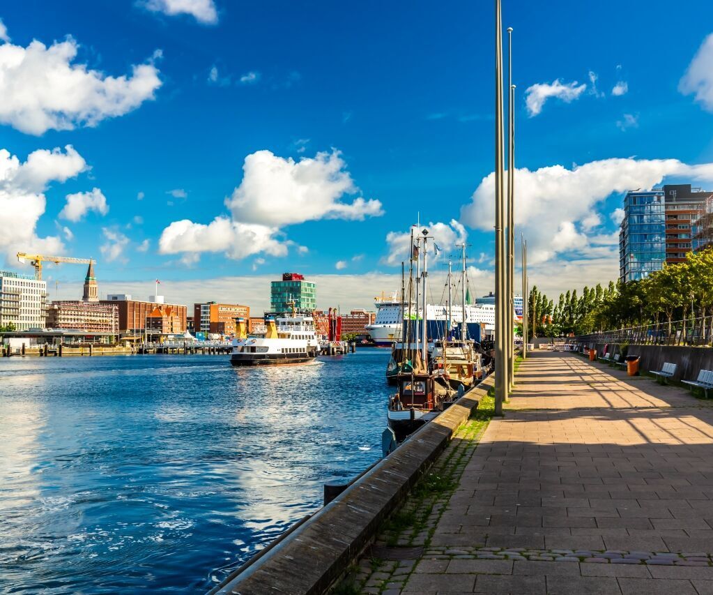Hafenpromenade bei Sonnenschein mit ruhigem Wasser, modernen Gebäuden im Hintergrund und Spazierweg am Wasser, der zur entspannten Erkundung des urbanen Hafengeländes einlädt.