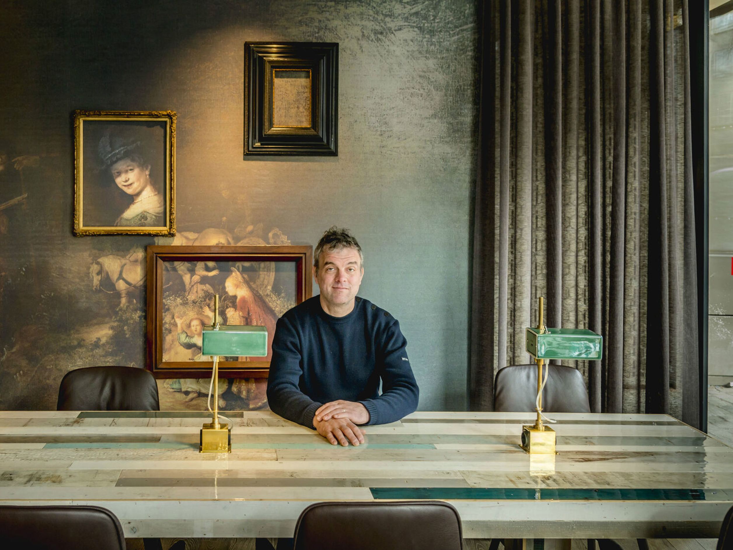 Mann sitzt in modern gemütlich eingerichteter Lounge mit großem Marmor-Tisch, Retro-Lampen und stilvollen Gemälden an der Wand.