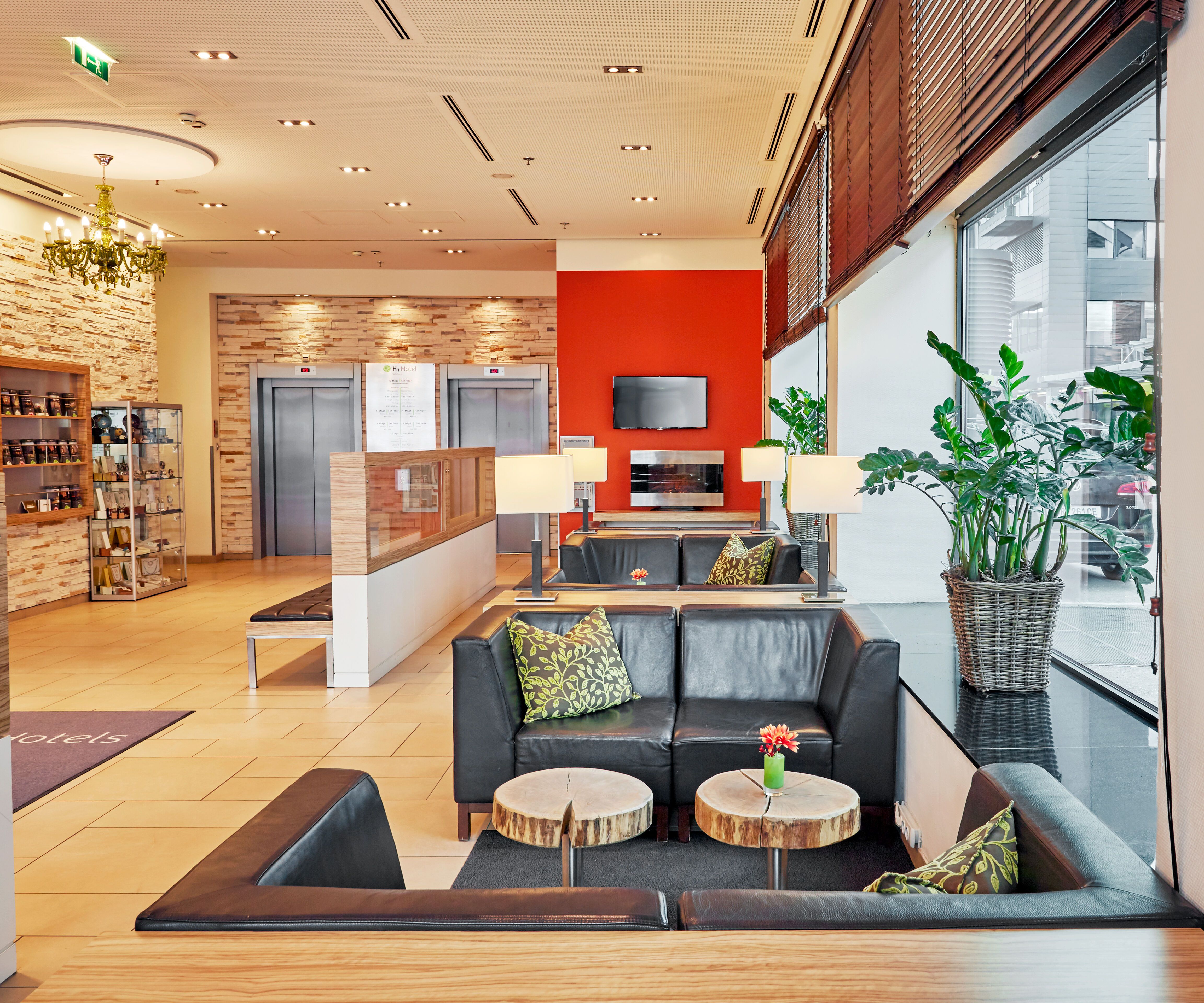 Moderne Hotellobby mit bequemen schwarzen Ledersofas, kleinen Holztischen, großen Fenstern für Tageslicht und grünen Pflanzen, die eine freundliche und einladende Atmosphäre schaffen. Warme Beleuchtung und dezente Dekoration sorgen für Wohlfühlambiente.