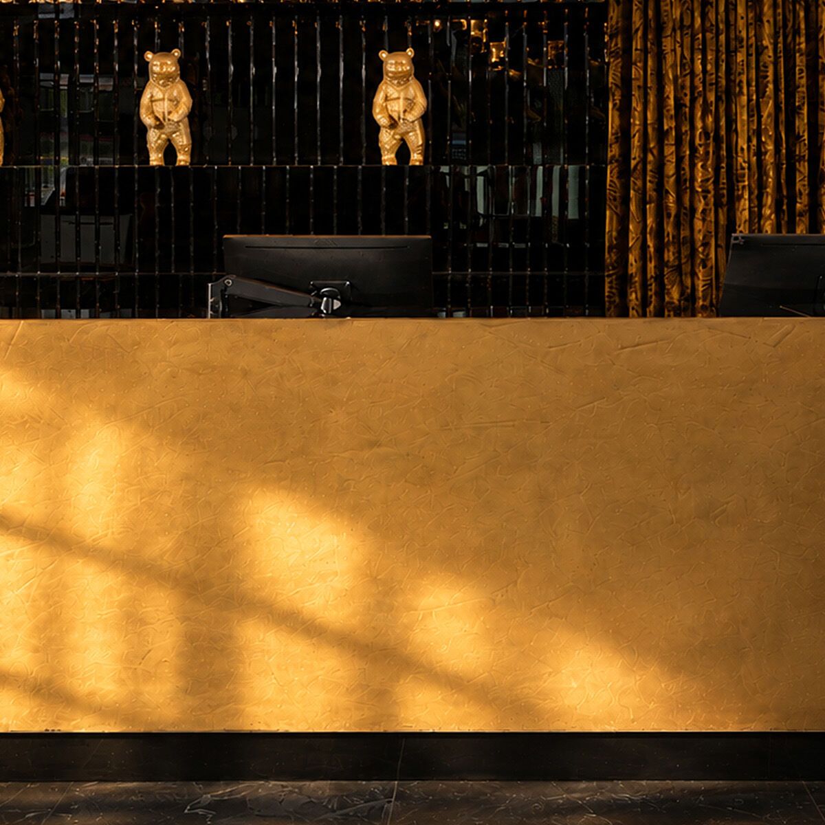 Elegante Hotelrezeption mit goldfarbenem Empfangstresen, schwarzem Hintergrund und stilvoller Beleuchtung. Drei goldene Bärenfiguren als dekoratives Highlight. Ambiente vermittelt Luxus und Wärme.
