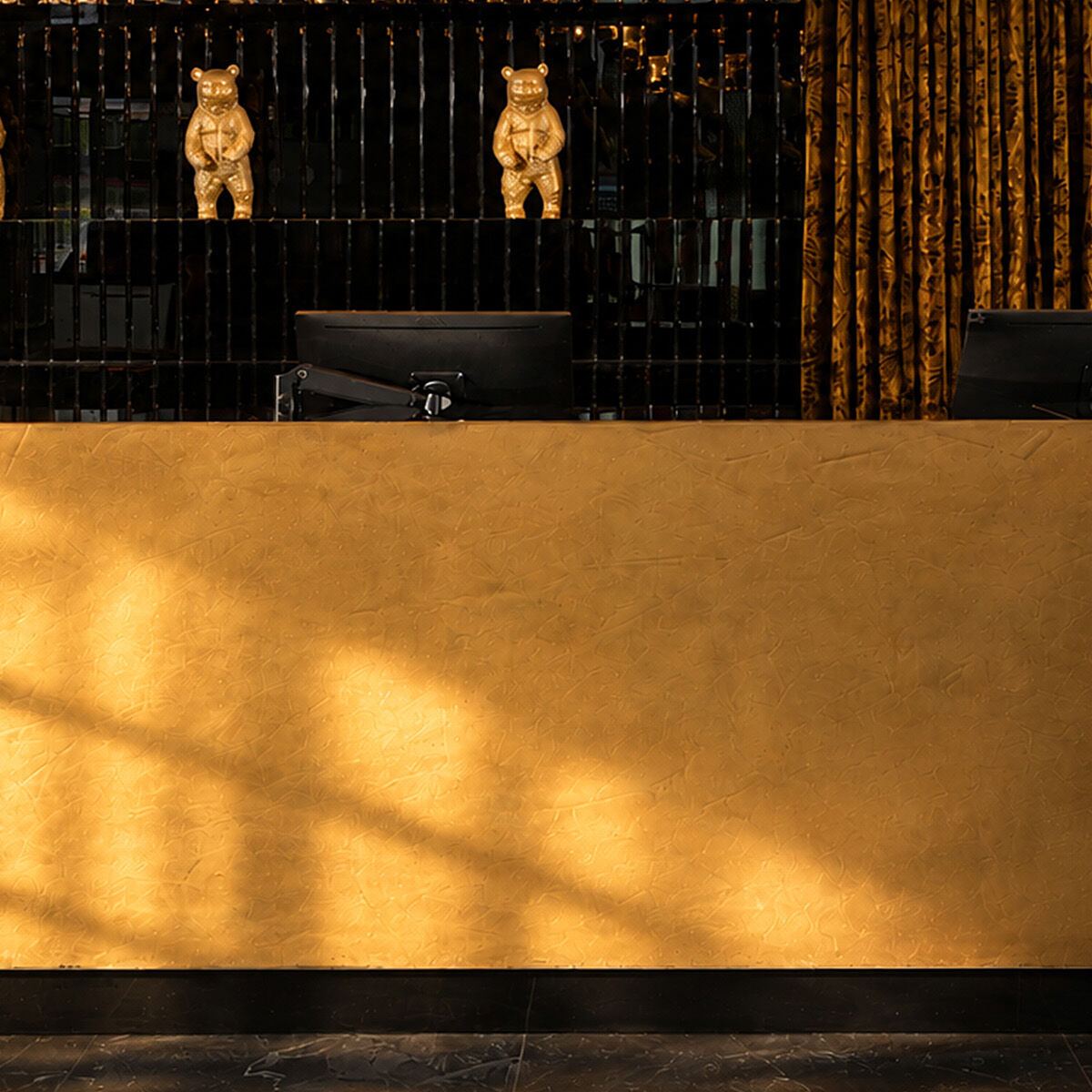 Elegante Hotelrezeption mit goldfarbenem Empfangstresen, schwarzem Hintergrund und stilvoller Beleuchtung. Drei goldene Bärenfiguren als dekoratives Highlight. Ambiente vermittelt Luxus und Wärme.