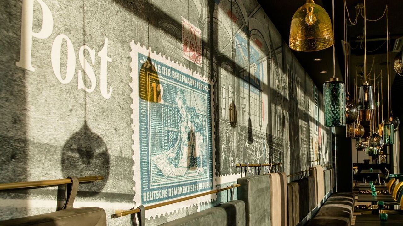 Lichtdurchfluteter Hotelbarbereich mit komfortabler Sitzbank und farblich kontrastierenden Stühlen, stilvoll dekoriert mit großen Briefmarkenmotiven an der Wand