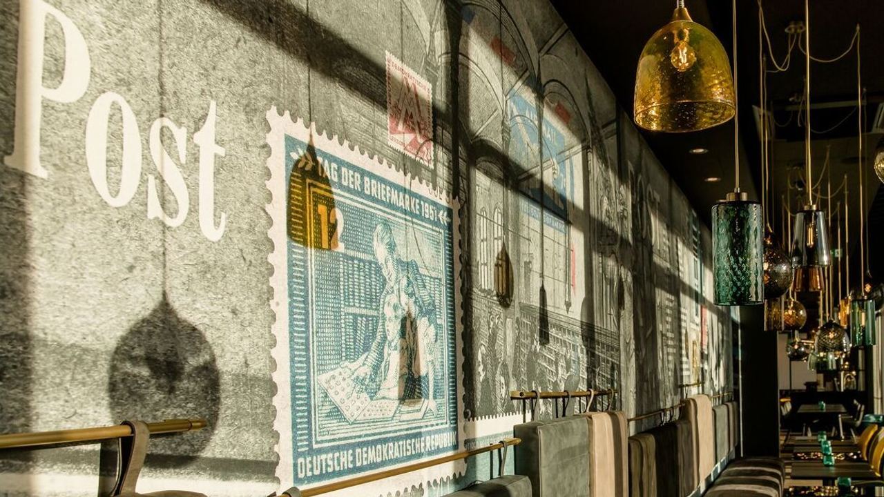 Lichtdurchfluteter Hotelbarbereich mit komfortabler Sitzbank und farblich kontrastierenden Stühlen, stilvoll dekoriert mit großen Briefmarkenmotiven an der Wand