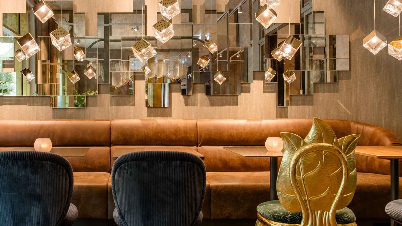 Eleganter Sitzbereich mit braunem Ledersofa, modernen schwarzen Stühlen und auffälligen goldfarbenen Stühlen, beleuchtet von warmen Hängelampen vor einer kunstvollen, spiegelnden Wanddekoration.