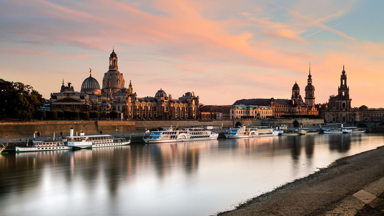 Panoramablick auf die historische Altstadt von Dresden mit der Elbe im Vordergrund, sanft beleuchtet vom warmen Licht des Sonnenuntergangs. Mehrere Ausflugsschiffe liegen am Flussufer, das ruhige Wasser spiegelt die malerische Skyline.