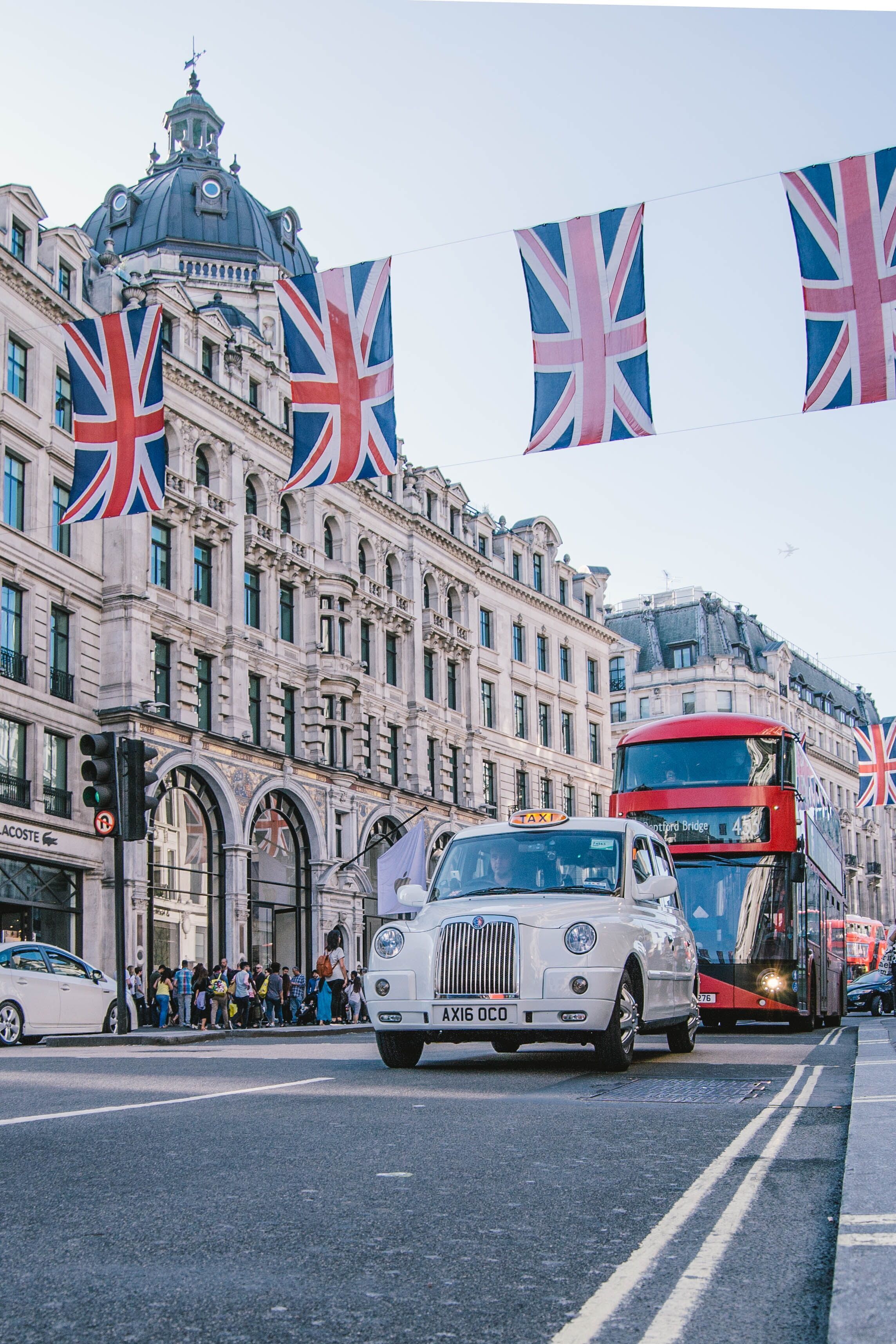 Straße in London mit britischen Flaggen über der Fahrbahn, traditionellen schwarzen Taxis und einem roten Doppeldeckerbus vor historischen Gebäuden im Stadtzentrum.