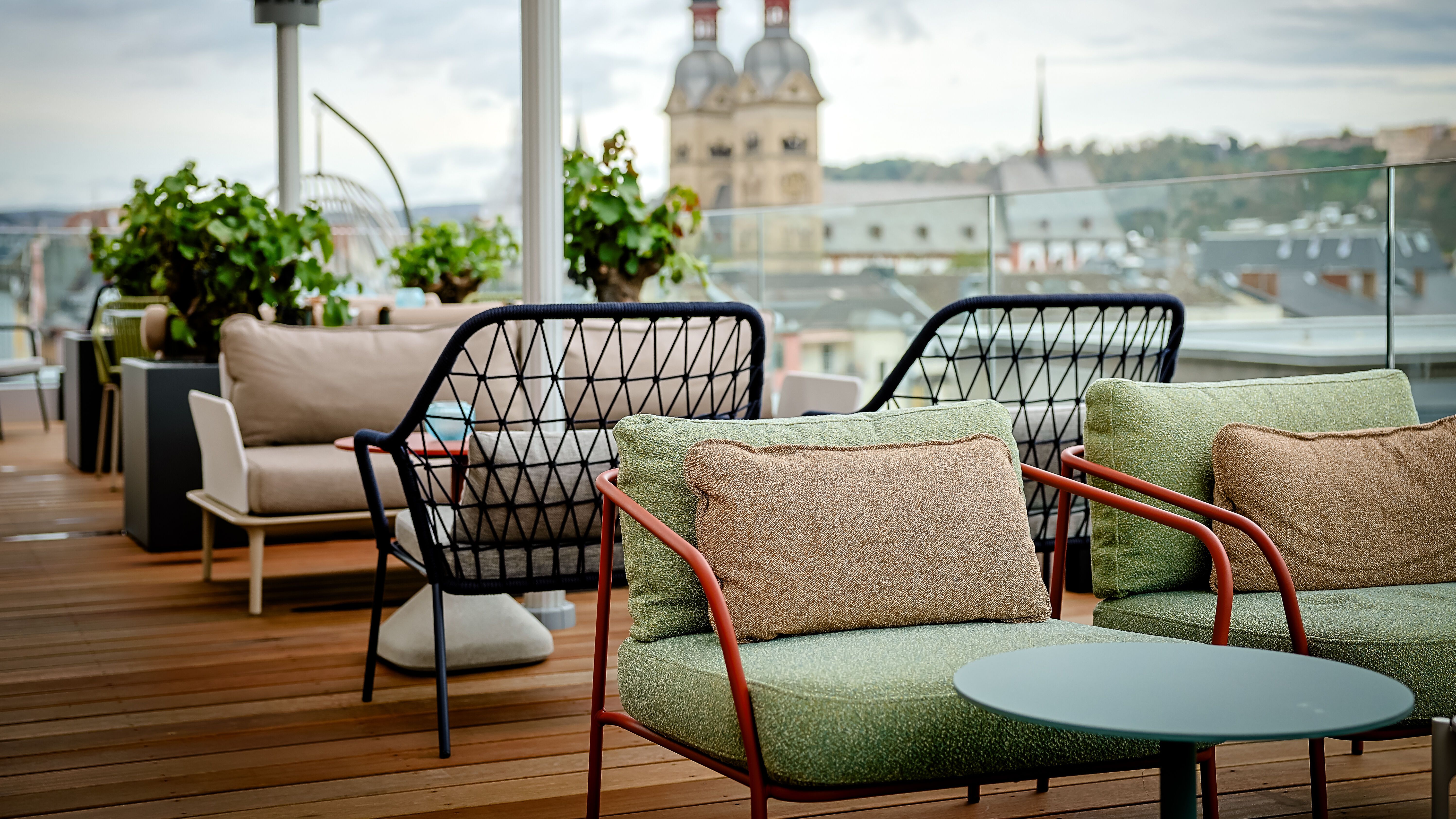 Gemütliche Dachterrassen-Lounge mit gepolsterten Sesseln und Tischen, umgeben von Pflanzen, mit Blick auf historische Kirchtürme in der Stadt. Ideal zum Entspannen und Genießen der Aussicht.