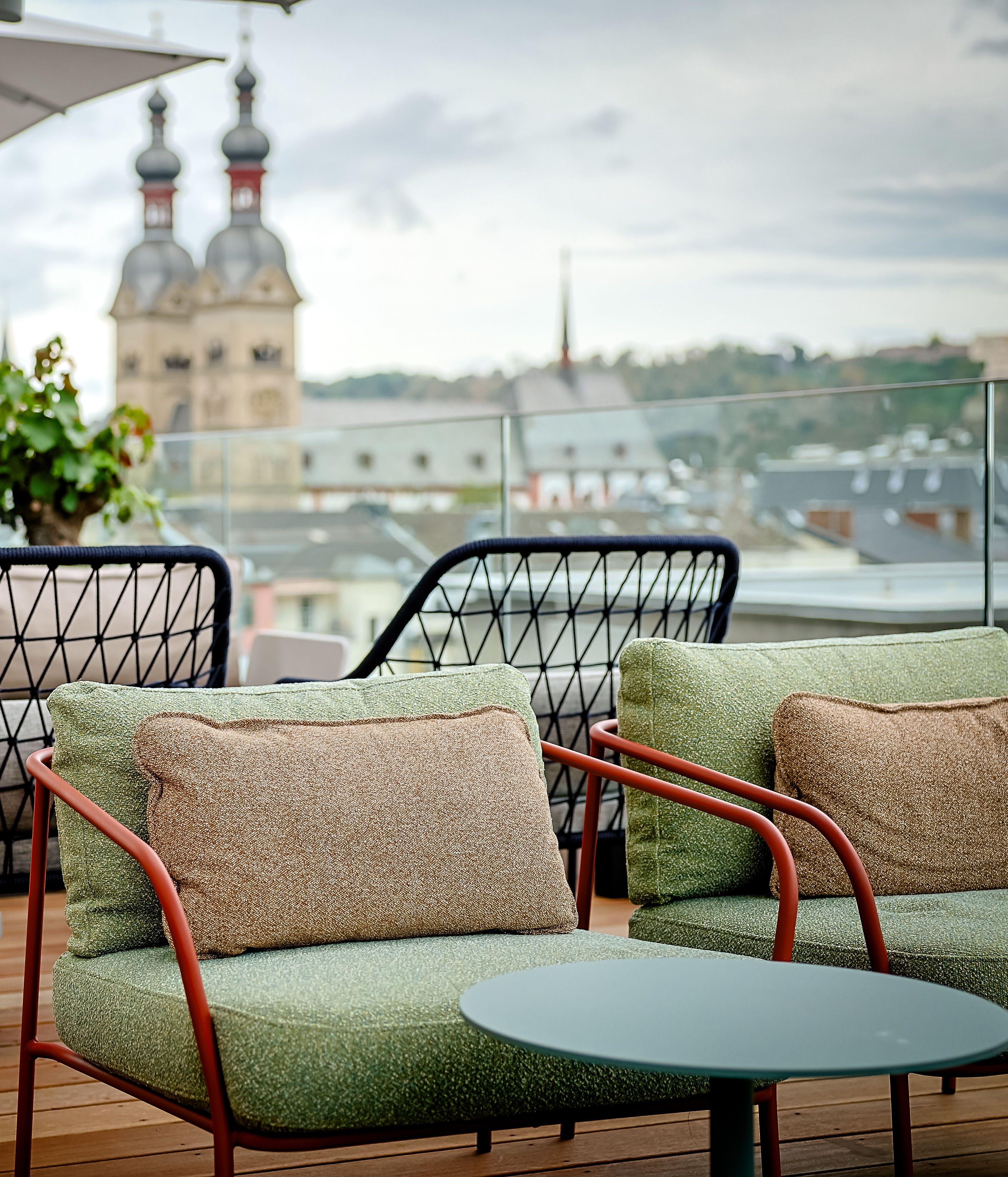 Gemütliche Dachterrassen-Lounge mit gepolsterten Sesseln und Tischen, umgeben von Pflanzen, mit Blick auf historische Kirchtürme in der Stadt. Ideal zum Entspannen und Genießen der Aussicht.