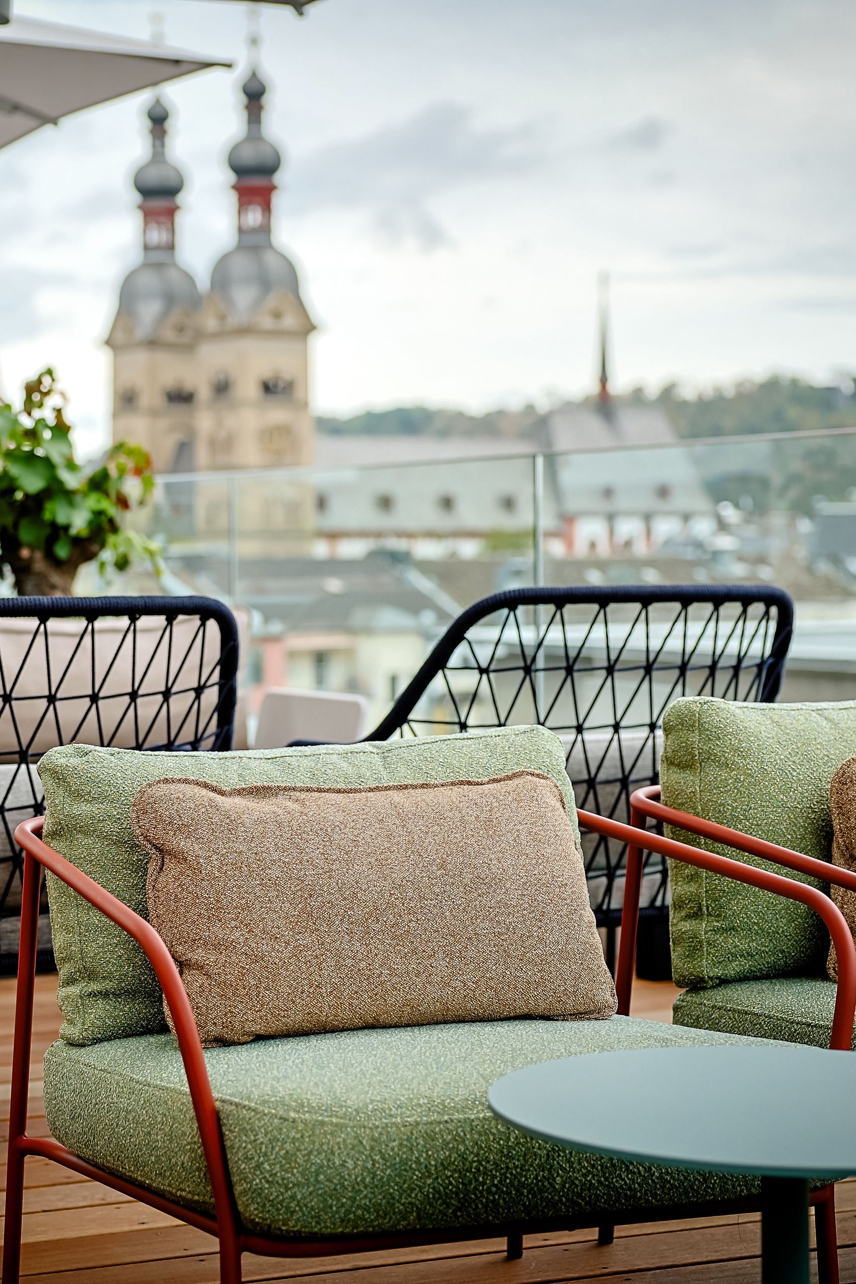 Gemütliche Dachterrassen-Lounge mit gepolsterten Sesseln und Tischen, umgeben von Pflanzen, mit Blick auf historische Kirchtürme in der Stadt. Ideal zum Entspannen und Genießen der Aussicht.