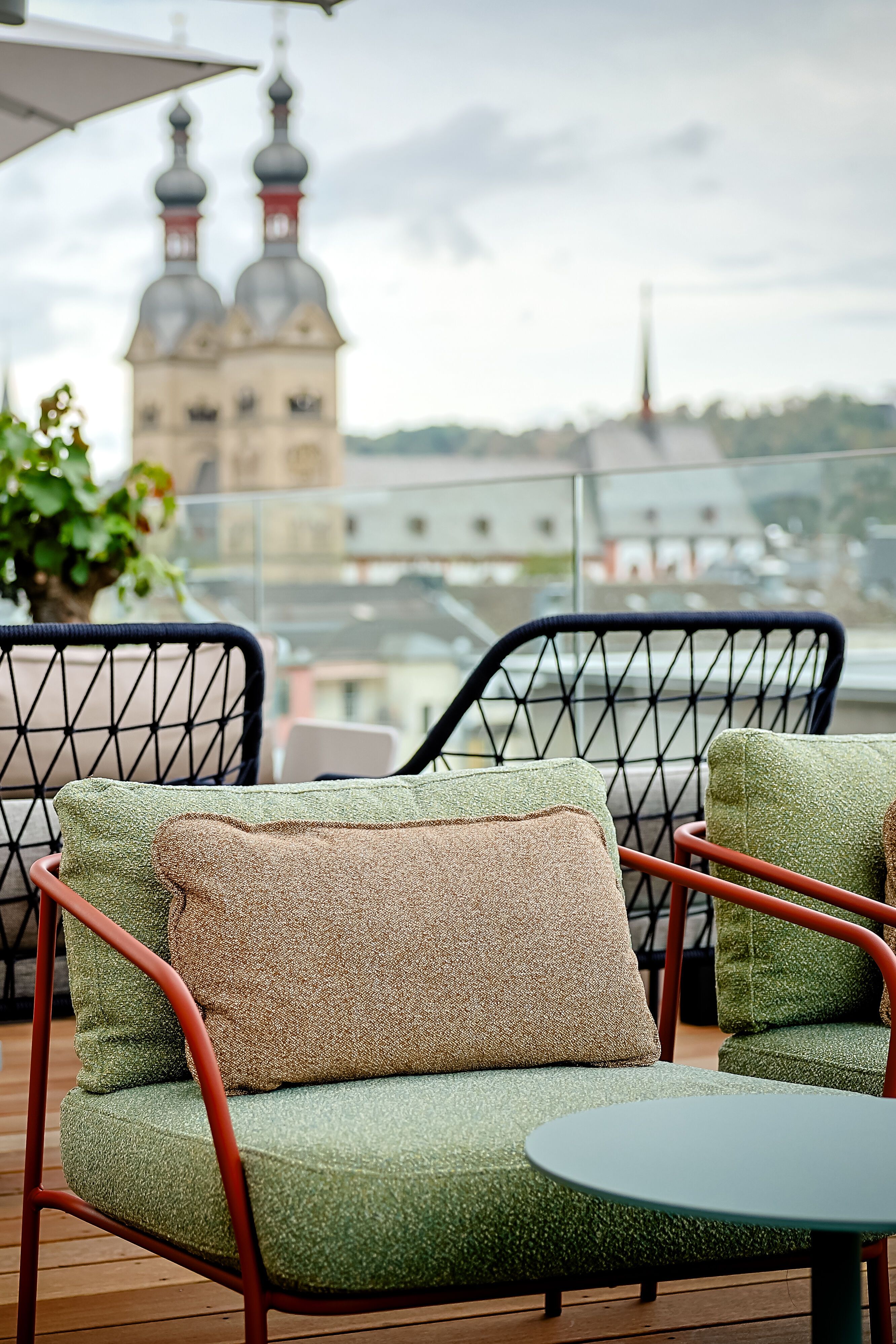 Gemütliche Dachterrassen-Lounge mit gepolsterten Sesseln und Tischen, umgeben von Pflanzen, mit Blick auf historische Kirchtürme in der Stadt. Ideal zum Entspannen und Genießen der Aussicht.