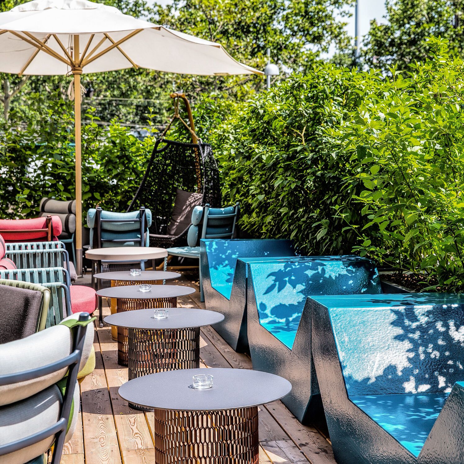 Motel One Außenlounge mit moderner Sitzmöblierung, blauen Kunststoffsofas, kleinen Tischen und Sonnenschirm, umgeben von üppigem Grün.