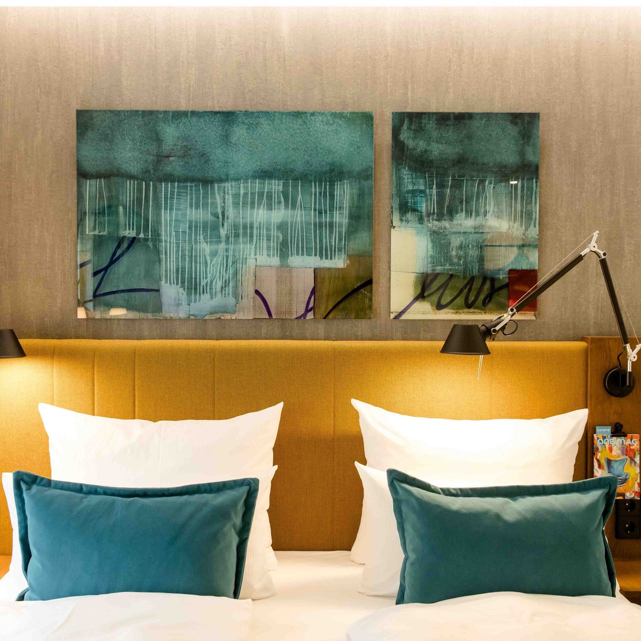 Motel One Hotelzimmer mit gemütlichem Doppelbett, gelbem Kopfteil, weißen Bettdecken und grünen Akzentkissen. An der Wand hängen abstrakte blaue Kunstwerke, dazu flexible Leselampen für angenehmes Licht.