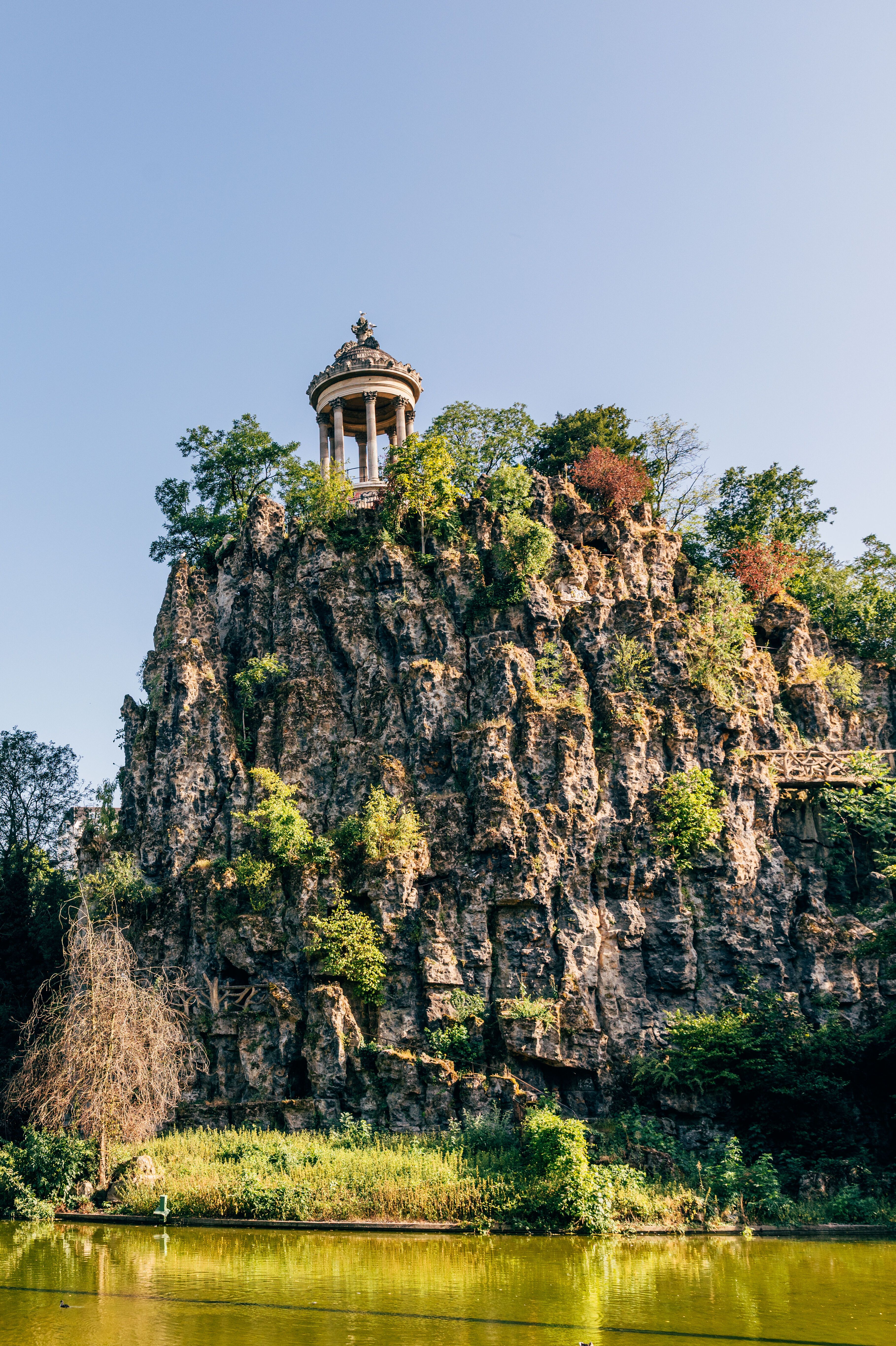 Blick auf einen felsigen Hügel mit einem Pavillon oben, umgeben von grünen Bäumen und einem ruhigen See im Vordergrund, bei klarem Himmel.