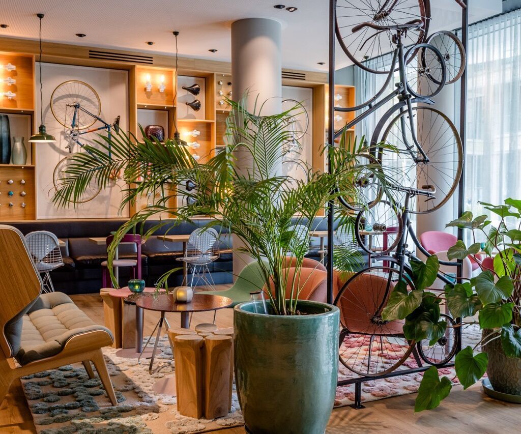 Helle Lounge mit modernen Sitzmöbeln, großen Pflanzen und dekorativ an der Wand und von der Decke hängenden Fahrrädern, die eine entspannte und stilvolle Atmosphäre schaffen.