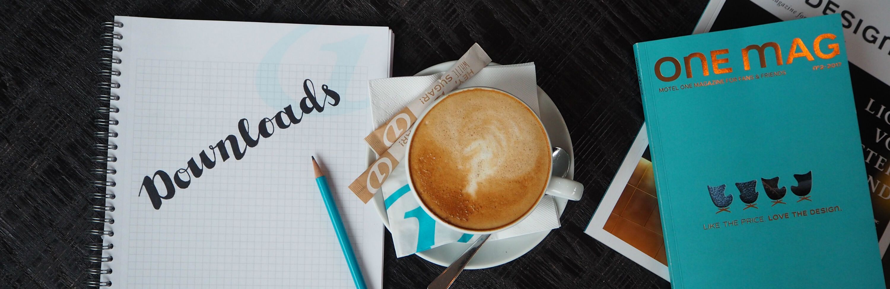 Notizblock mit dem Wort „Downloads“, daneben eine Tasse Kaffee mit Milchschaum, ein Bleistift und eine Ausgabe des Magazins „one MAG“.