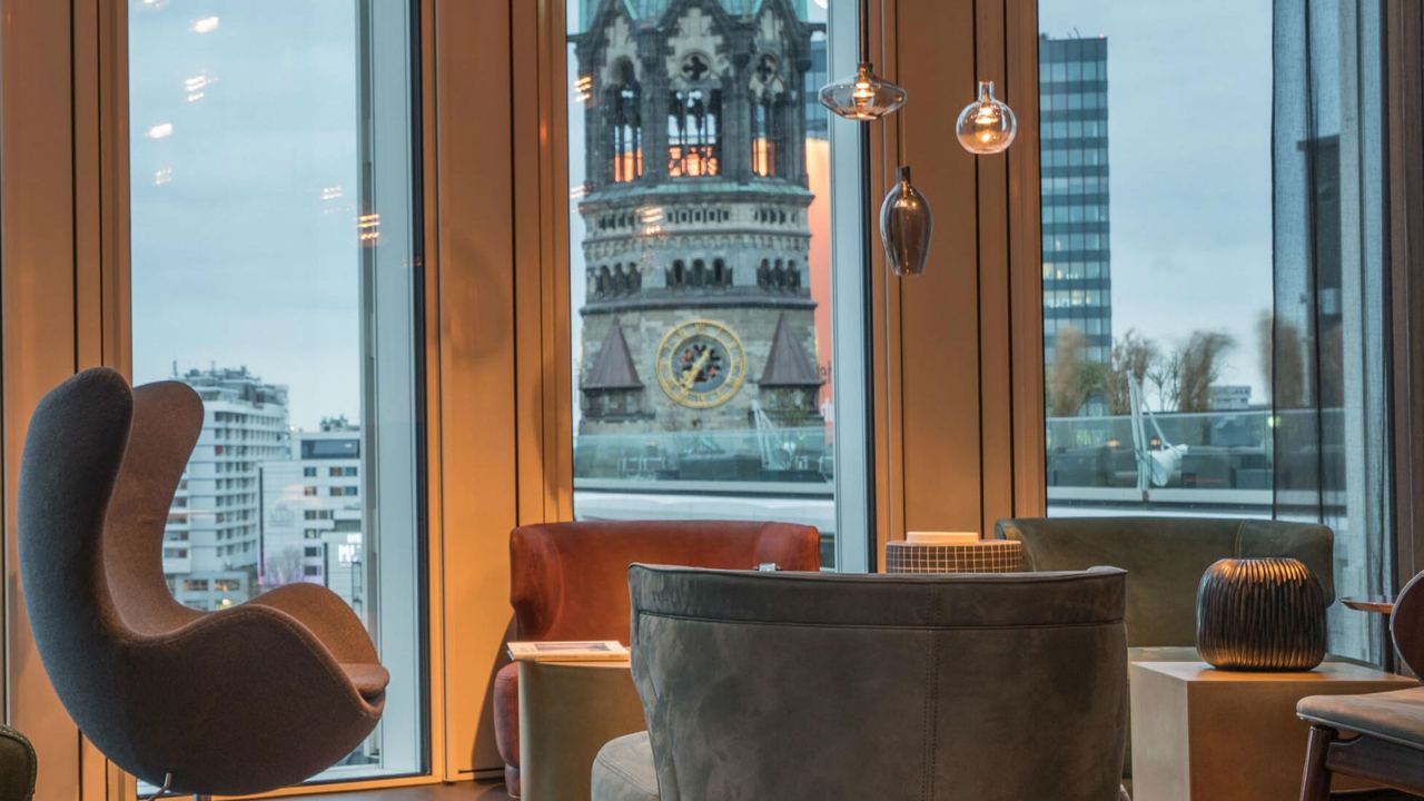 Gemütliche Sitzecke mit Sesseln und Hängeleuchten vor großen Fenstern, die einen Blick auf den historischen Turm der Kaiser-Wilhelm-Gedächtniskirche in Berlin bieten.
