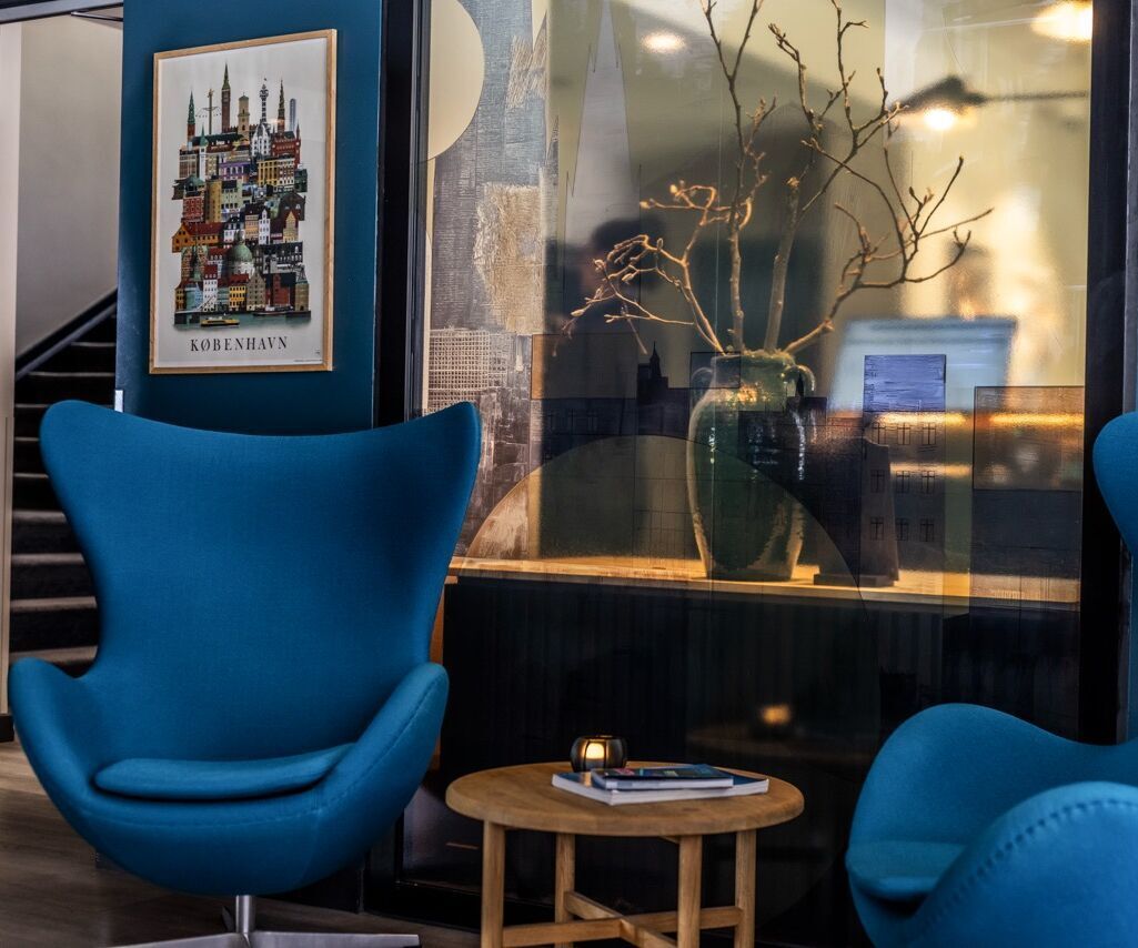Moderne Lounge-Ecke mit zwei komfortablen blauen Sesseln, einem runden Holztisch und dekorativem Pflanzenarrangement in Vase, stilvoll eingerahmt von künstlerischer Wandgestaltung.