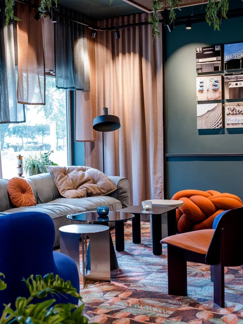 Gemütliche Lounge mit modernen, farbenfrohen Sesseln und Sofa, weichen Kissen, indirekter Beleuchtung und großen Fenstern, die Tageslicht hereinlassen. Pflanzen und Vorhänge schaffen eine entspannte, einladende Atmosphäre zum Verweilen.