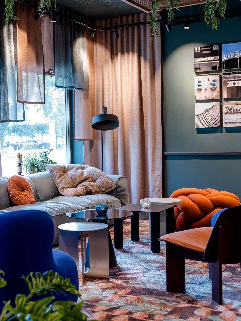 Gemütliche Lounge mit modernen, farbenfrohen Sesseln und Sofa, weichen Kissen, indirekter Beleuchtung und großen Fenstern, die Tageslicht hereinlassen. Pflanzen und Vorhänge schaffen eine entspannte, einladende Atmosphäre zum Verweilen.