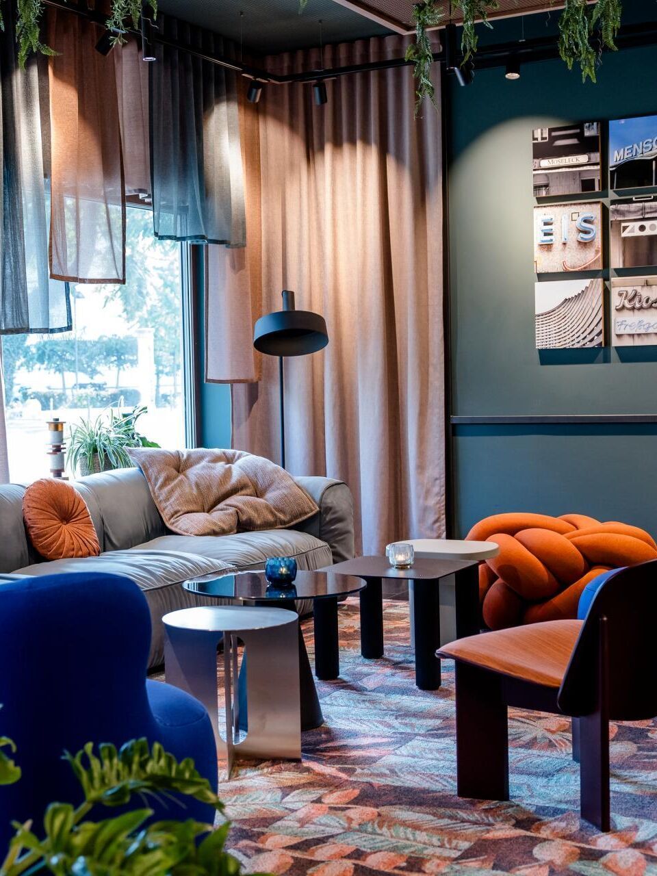 Gemütliche Lounge mit modernen, farbenfrohen Sesseln und Sofa, weichen Kissen, indirekter Beleuchtung und großen Fenstern, die Tageslicht hereinlassen. Pflanzen und Vorhänge schaffen eine entspannte, einladende Atmosphäre zum Verweilen.