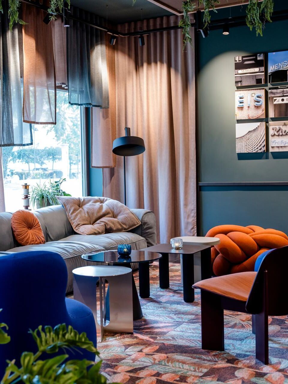 Gemütliche Lounge mit modernen, farbenfrohen Sesseln und Sofa, weichen Kissen, indirekter Beleuchtung und großen Fenstern, die Tageslicht hereinlassen. Pflanzen und Vorhänge schaffen eine entspannte, einladende Atmosphäre zum Verweilen.