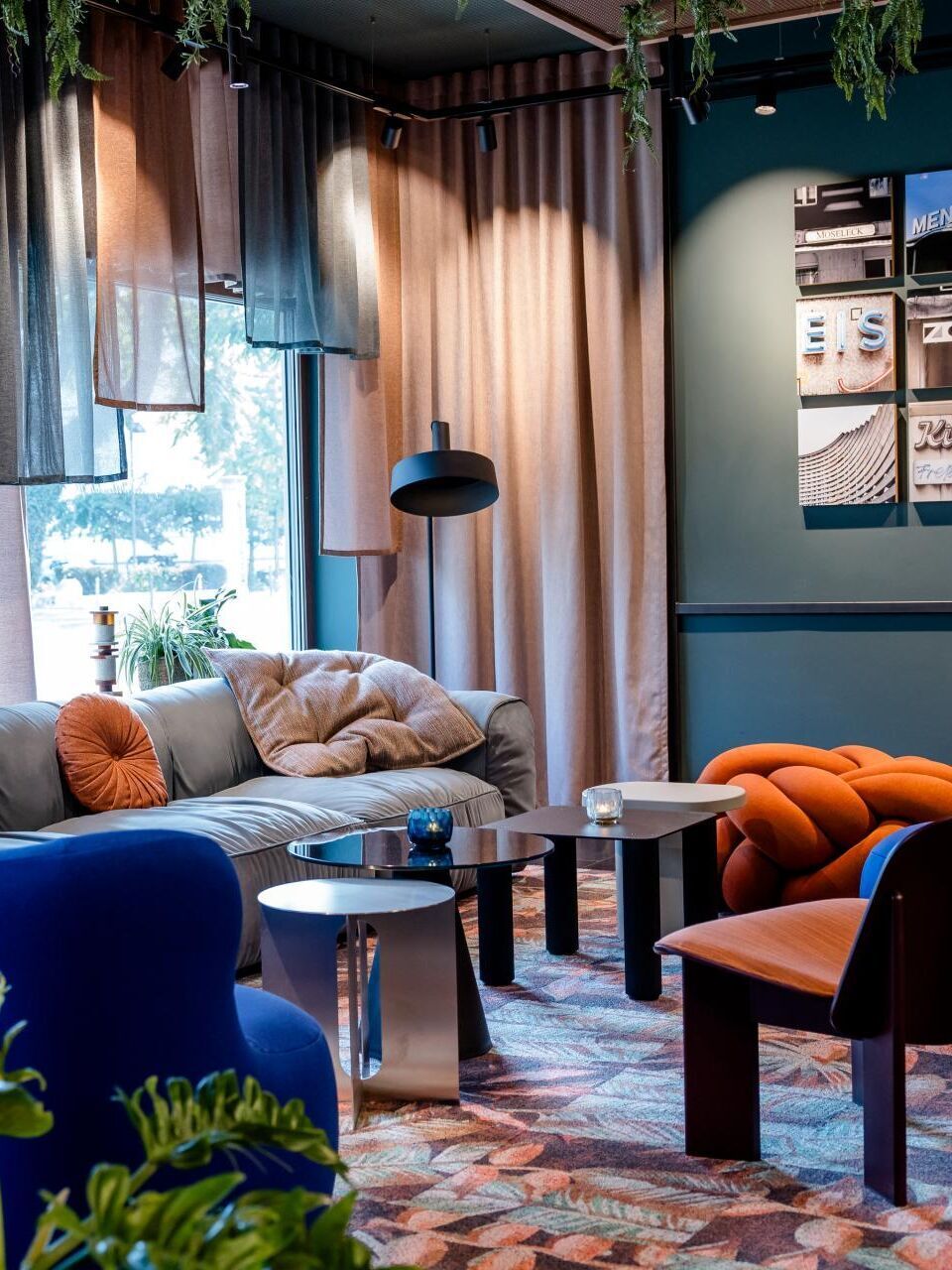 Gemütliche Lounge mit modernen, farbenfrohen Sesseln und Sofa, weichen Kissen, indirekter Beleuchtung und großen Fenstern, die Tageslicht hereinlassen. Pflanzen und Vorhänge schaffen eine entspannte, einladende Atmosphäre zum Verweilen.