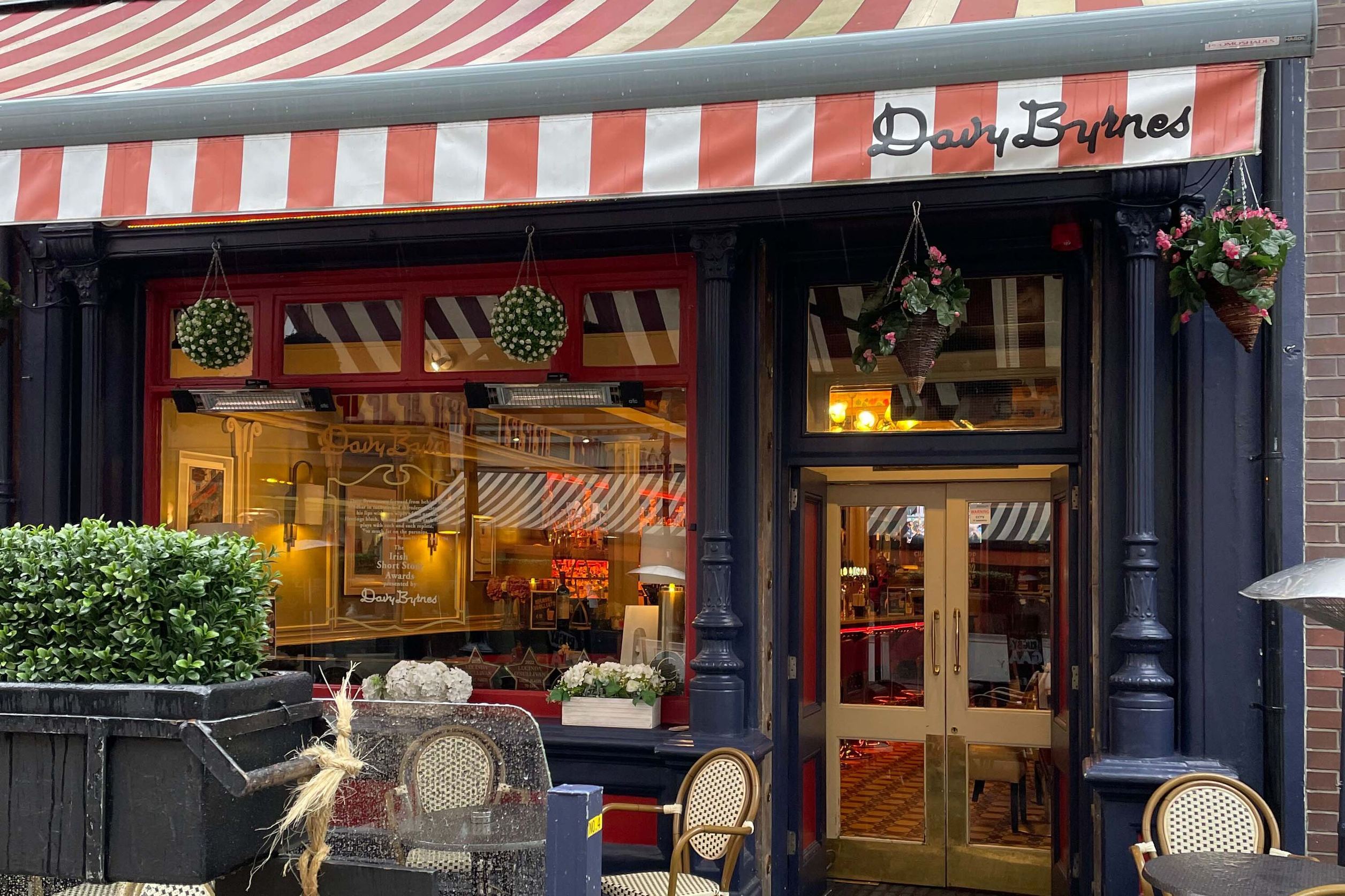 Außenansicht eines gemütlichen Cafés mit rot-weiß gestreiftem Sonnenschutz, Blumenarrangements und Sitzgelegenheiten im Freien, die zum Verweilen einladen.