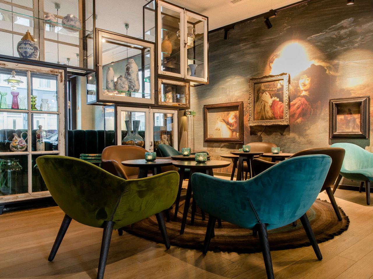 Gemütlicher Lounge-Bereich mit samtigen Sesseln in Grün und Blau, runden Tischen und stilvoller Wandbeleuchtung. An der Wand hängen klassische Gemälde, die dem Raum eine elegante und entspannte Atmosphäre verleihen. Große Fenster bringen Tageslicht herein und spiegelnde Vitrinen zeigen dekorative Objekte.