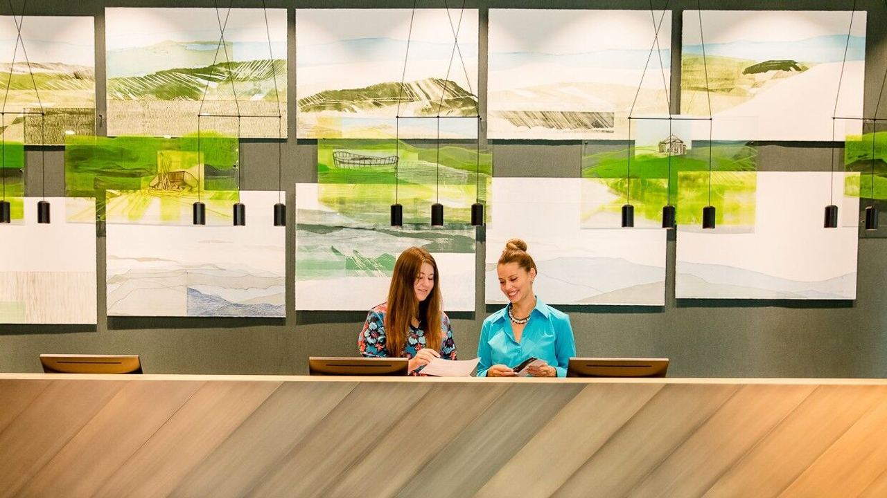 Zwei freundliche Mitarbeiterinnen am Hotelempfang besprechen gemeinsam Unterlagen, vor einer wand mit modernen Landschaftsbildern.