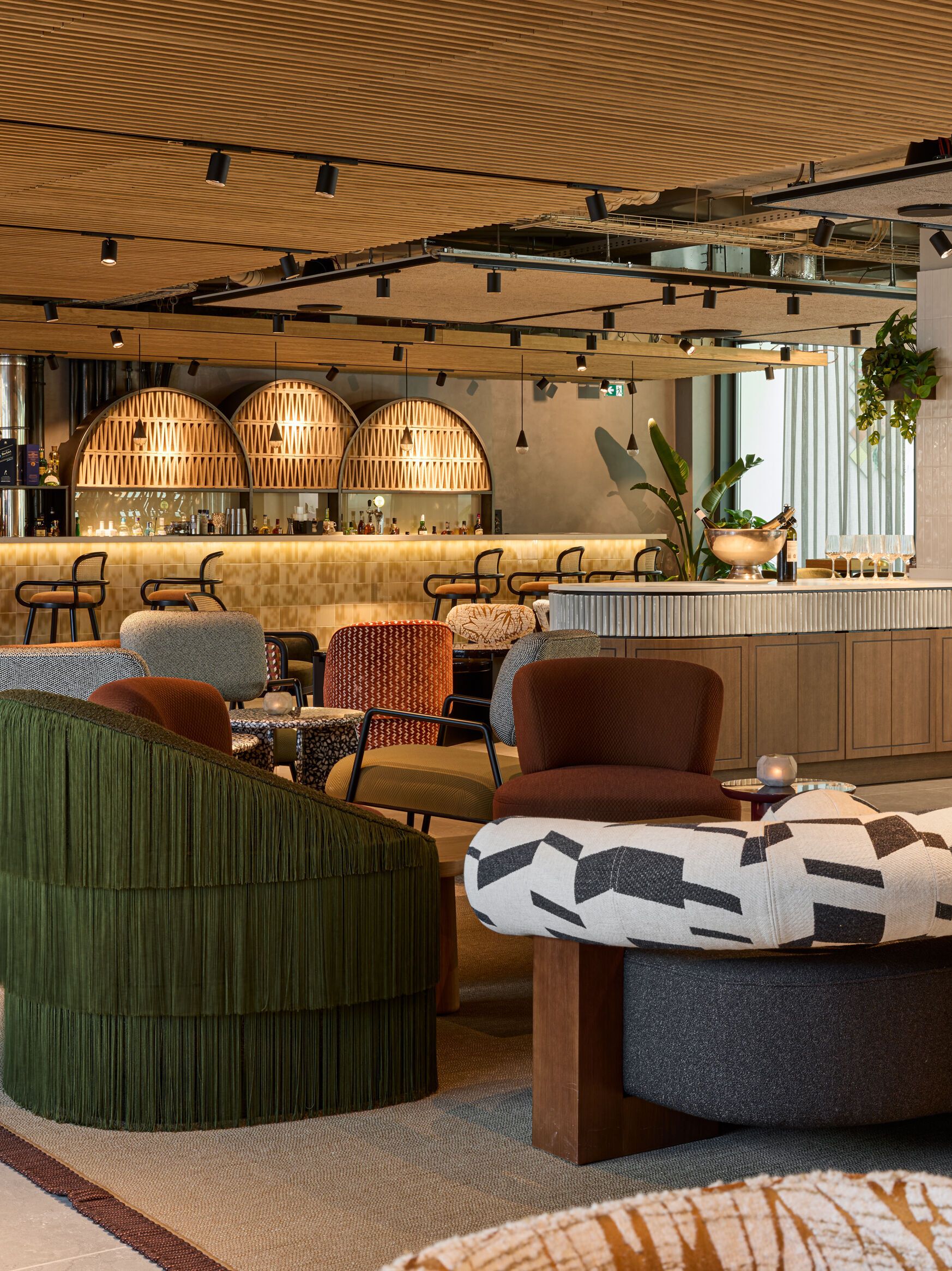 Moderne Hotelbar mit stilvollen Sitzgelegenheiten, warmem Holzdekor und atmosphärischer Beleuchtung. Gemütliche Lounge-Möbel laden zu entspannten Gesprächen bei einem Getränk ein, umgeben von grünen Pflanzen und elegantem Interieur.